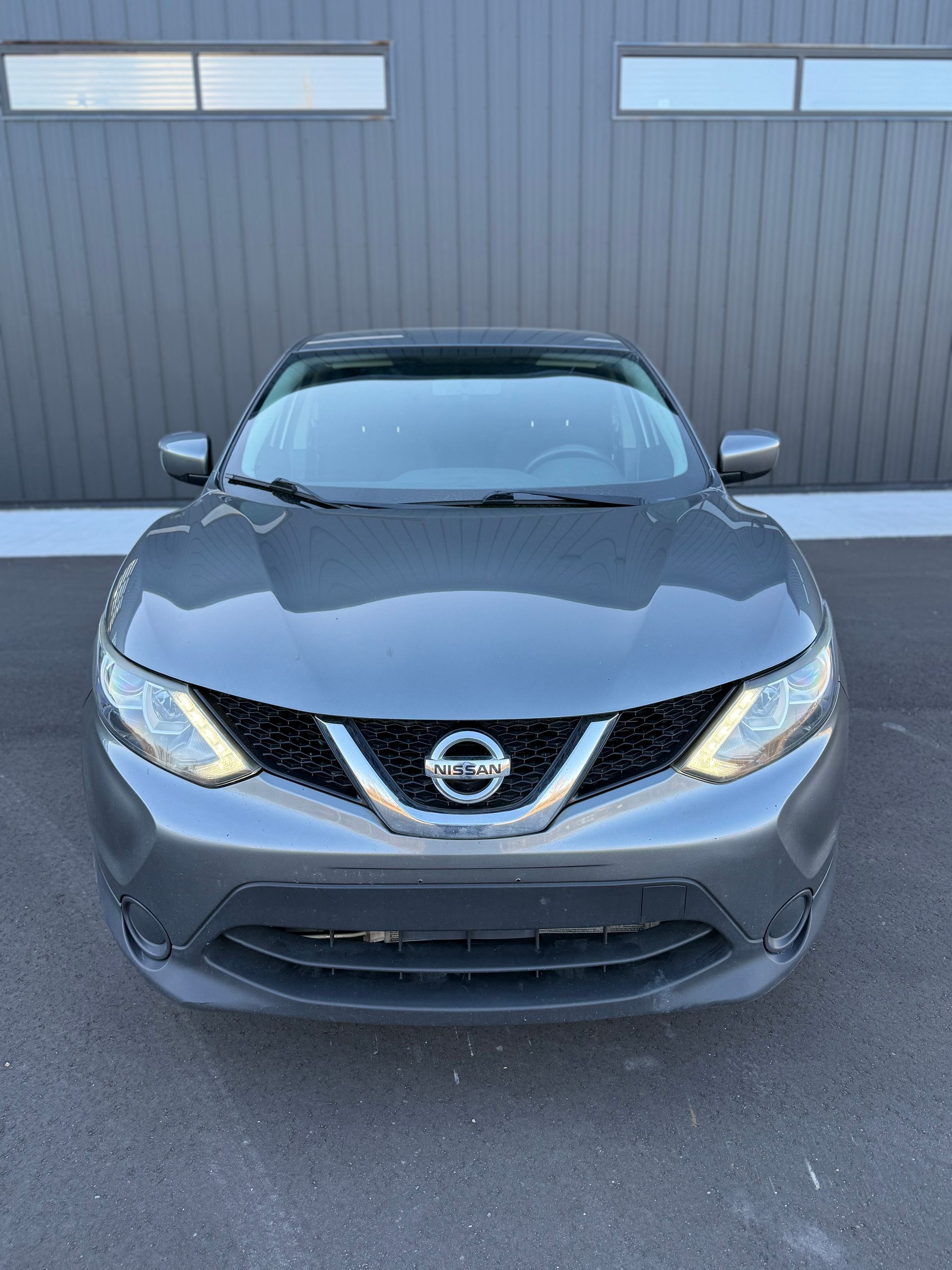 A gray Nissan Rogue parked on an asphalt lot in front of a dark-paneled industrial building.