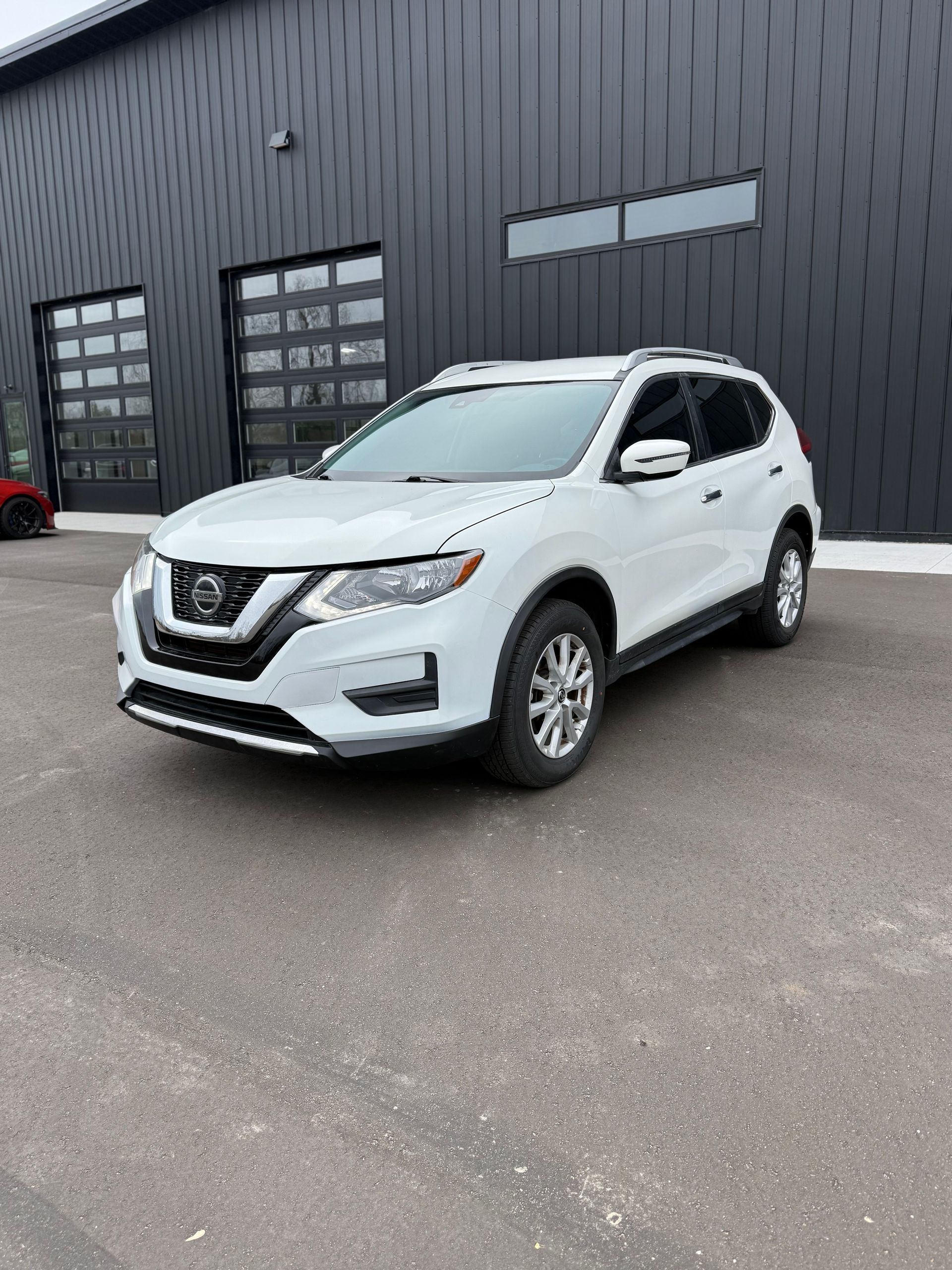 A white Nissan Rogue SUV parked on an asphalt lot in front of a dark, corrugated metal building.