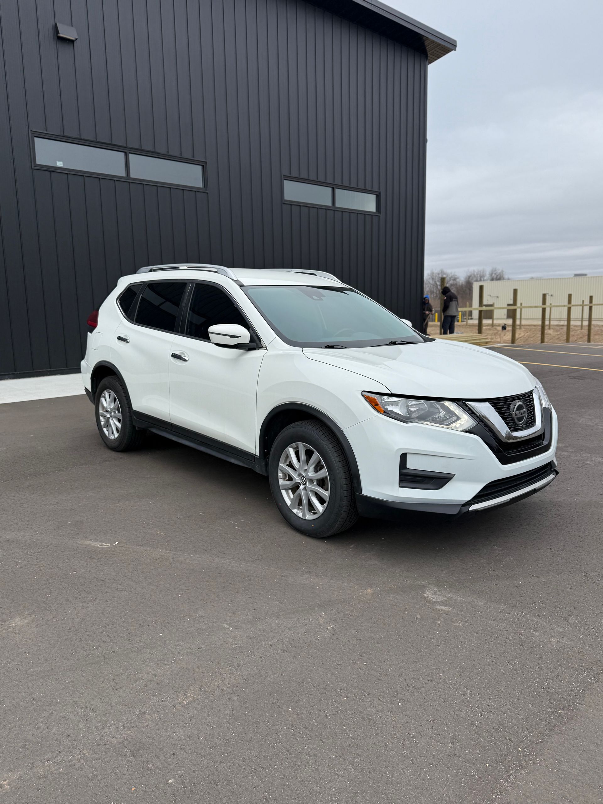 A white Nissan Rogue SUV parked on an asphalt lot in front of a dark, corrugated metal building.