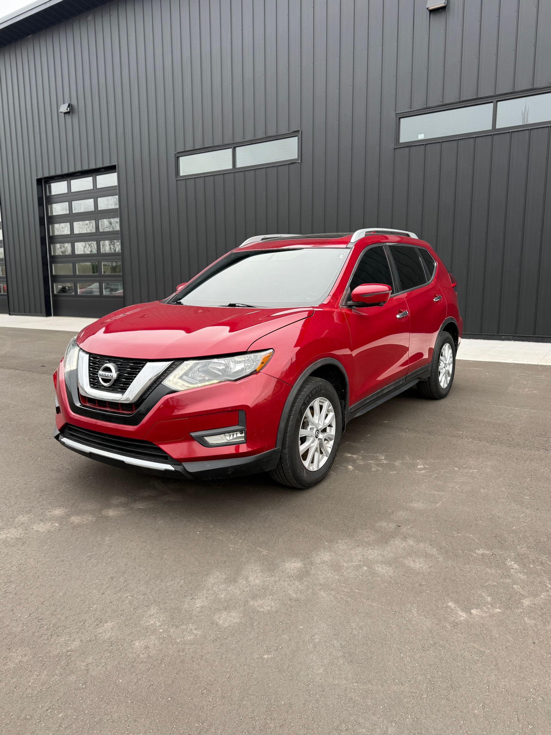 A red Nissan Rogue SUV parked on an asphalt lot in front of a black metal building.