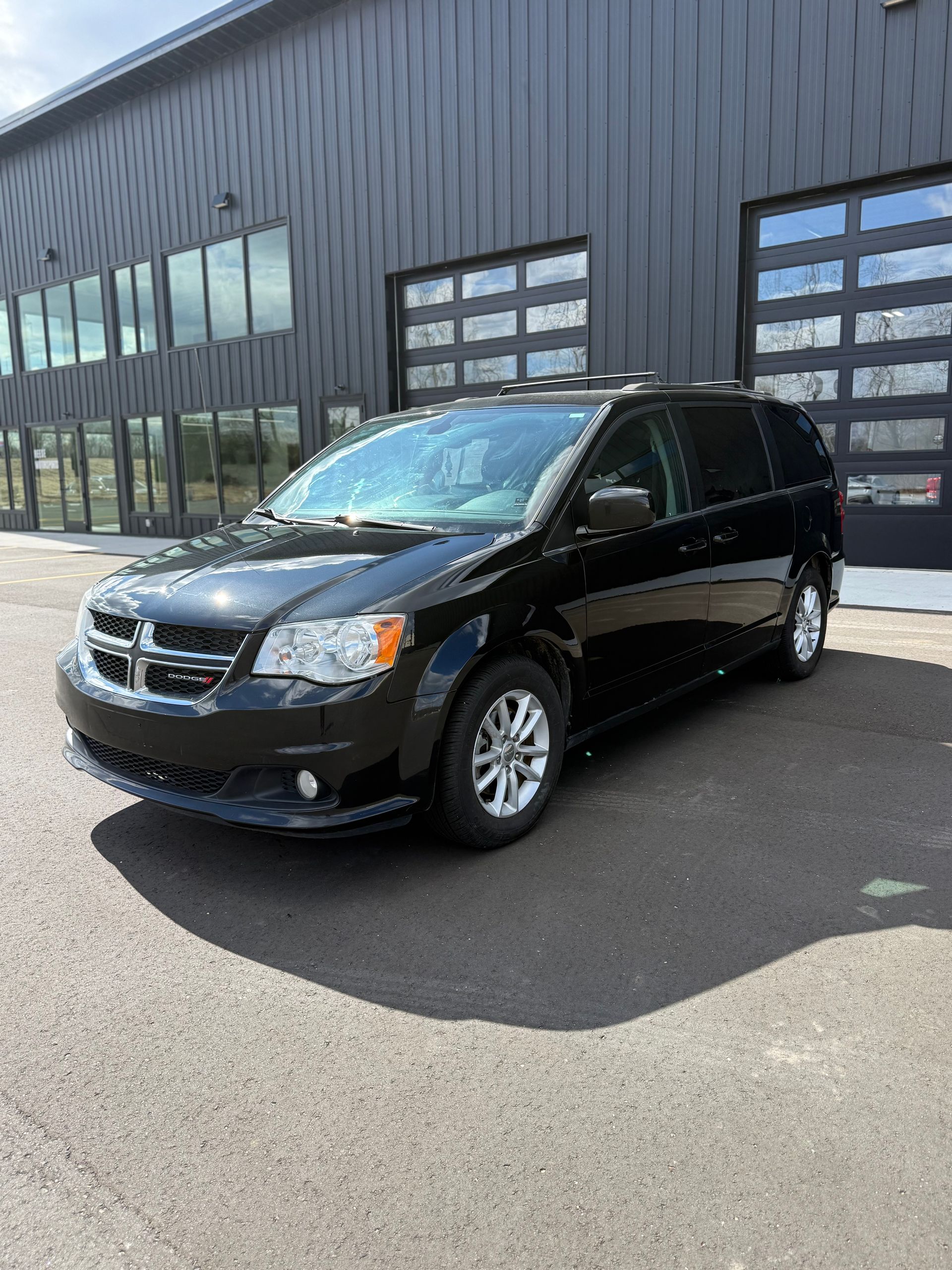 A black Dodge Grand Caravan parked in front of a modern building with large glass garage doors.