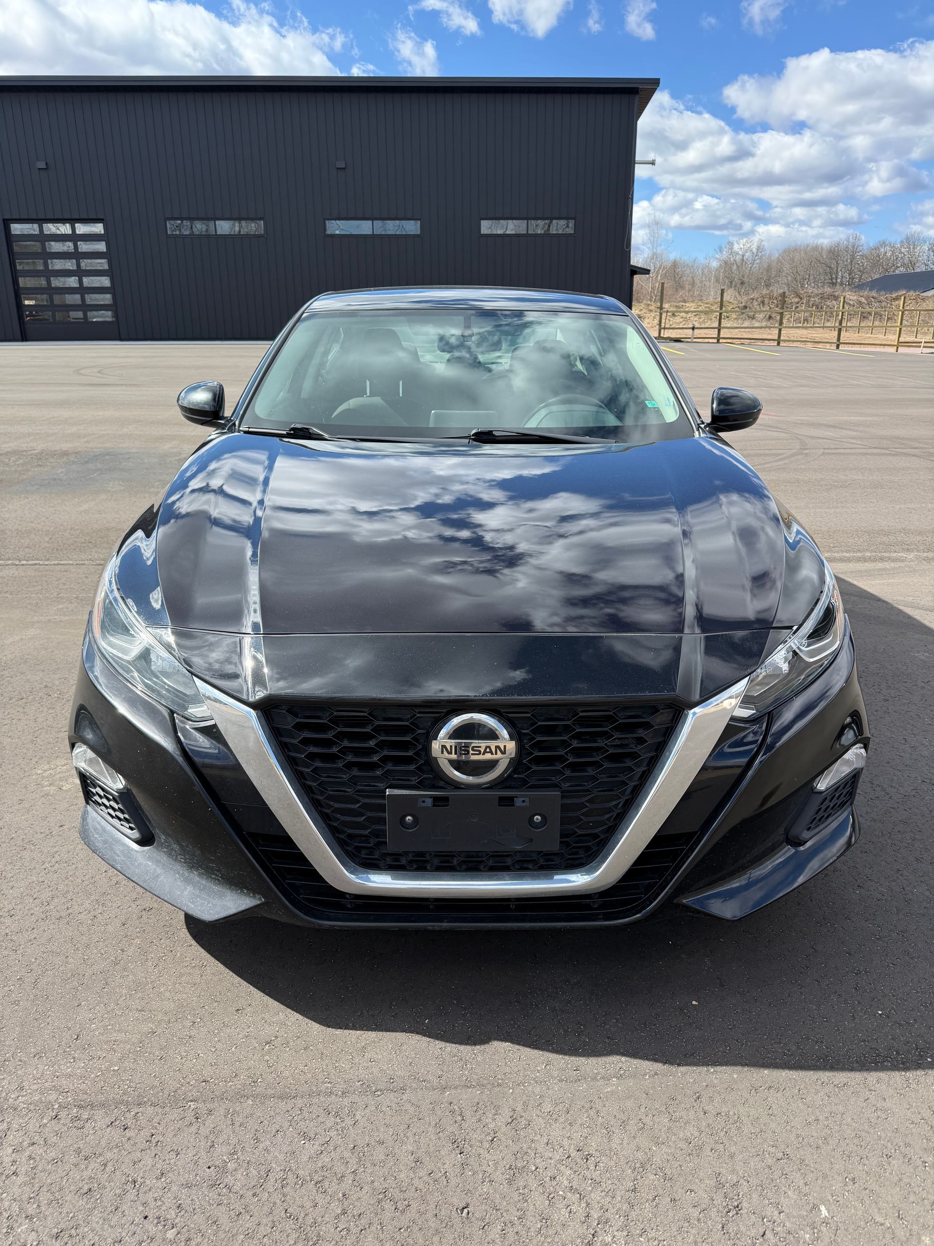 A black Nissan Altima parked in front of a dark gray building under a blue, cloudy sky.