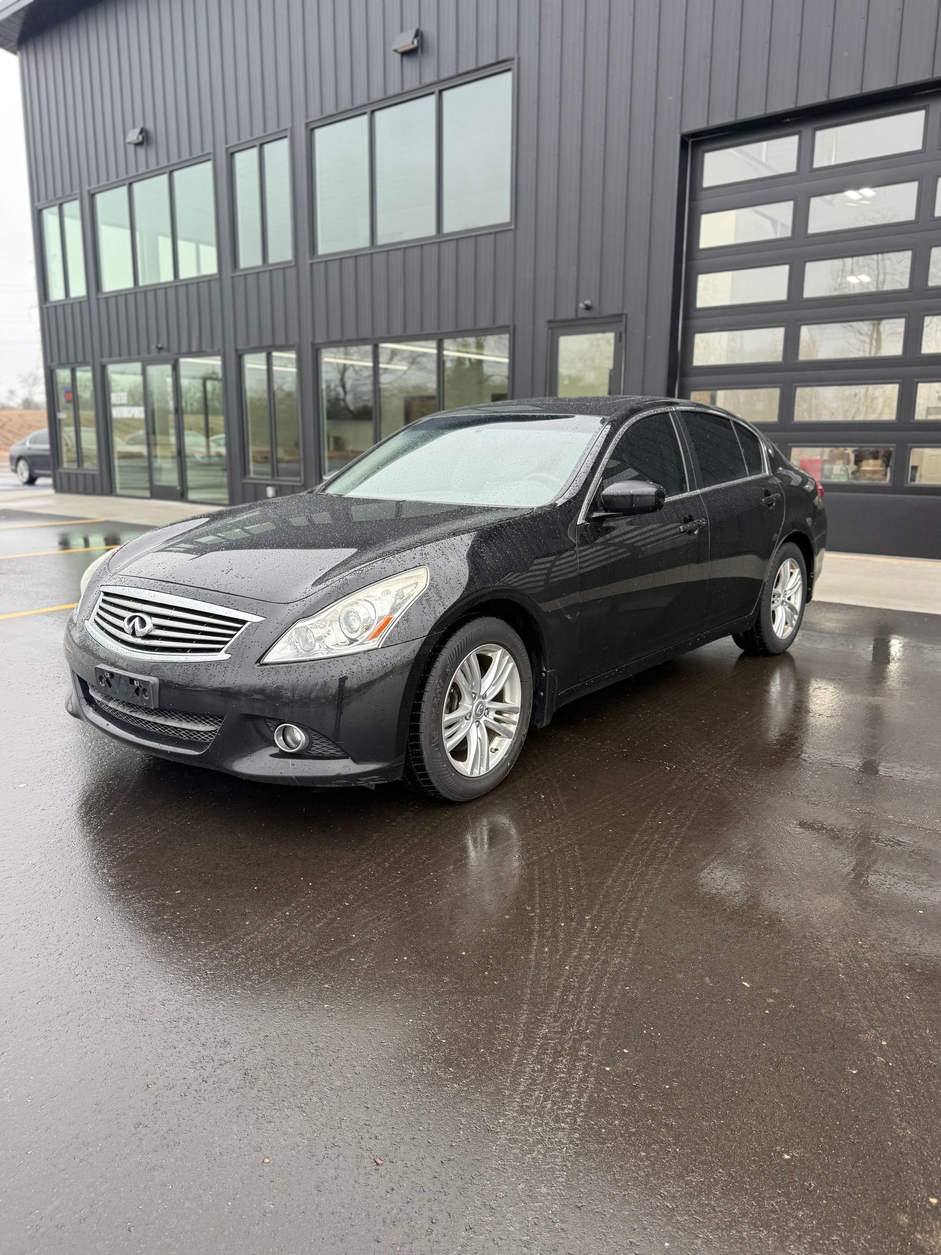 A dark gray Infiniti G sedan parked on a wet asphalt surface in front of a modern dark-paneled industrial building.