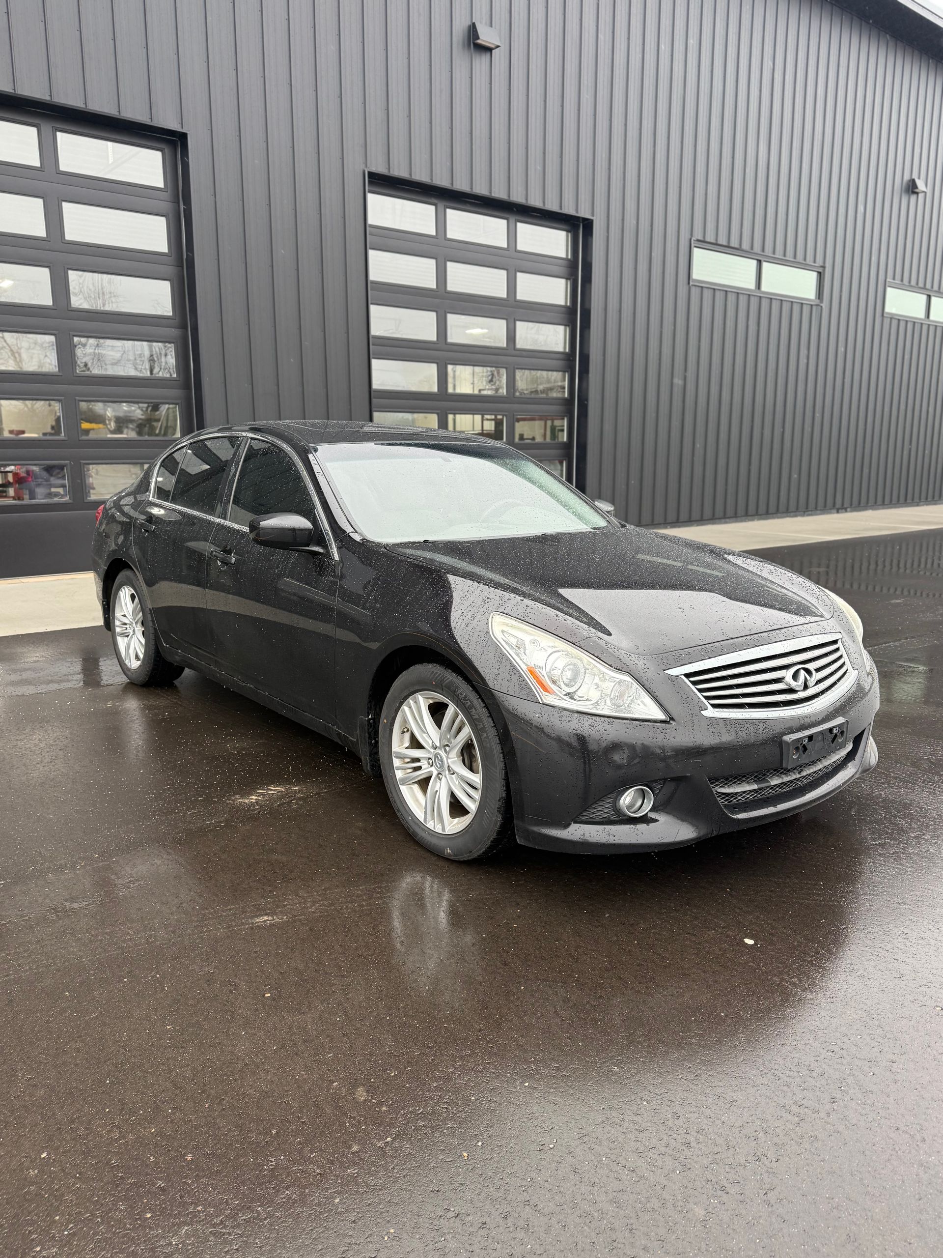 A dark gray Infiniti G sedan parked on a wet asphalt surface in front of a modern dark-paneled industrial building.