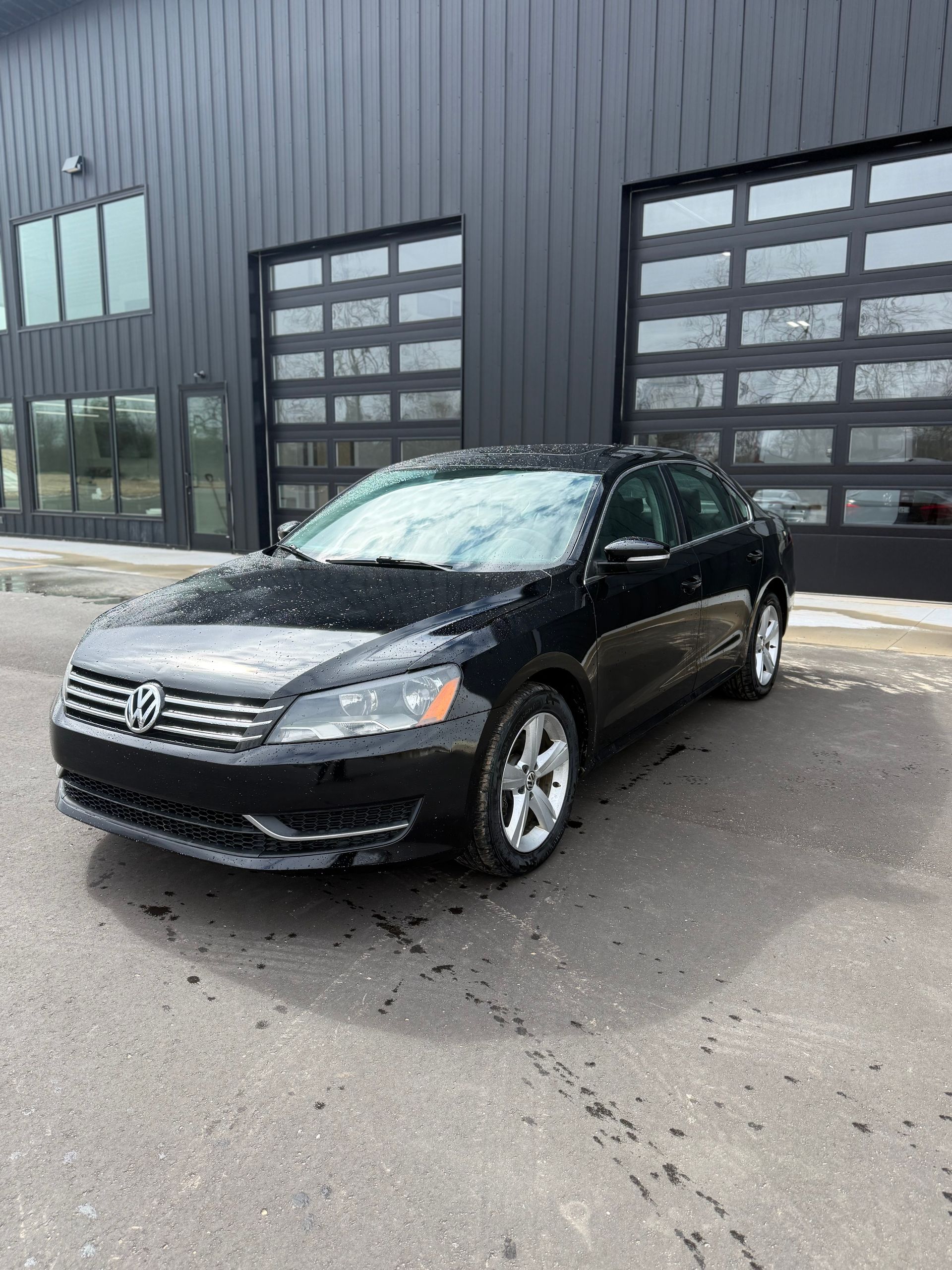 A black Volkswagen Passat sedan parked on a grey asphalt lot in front of a dark metal building with glass garage doors.