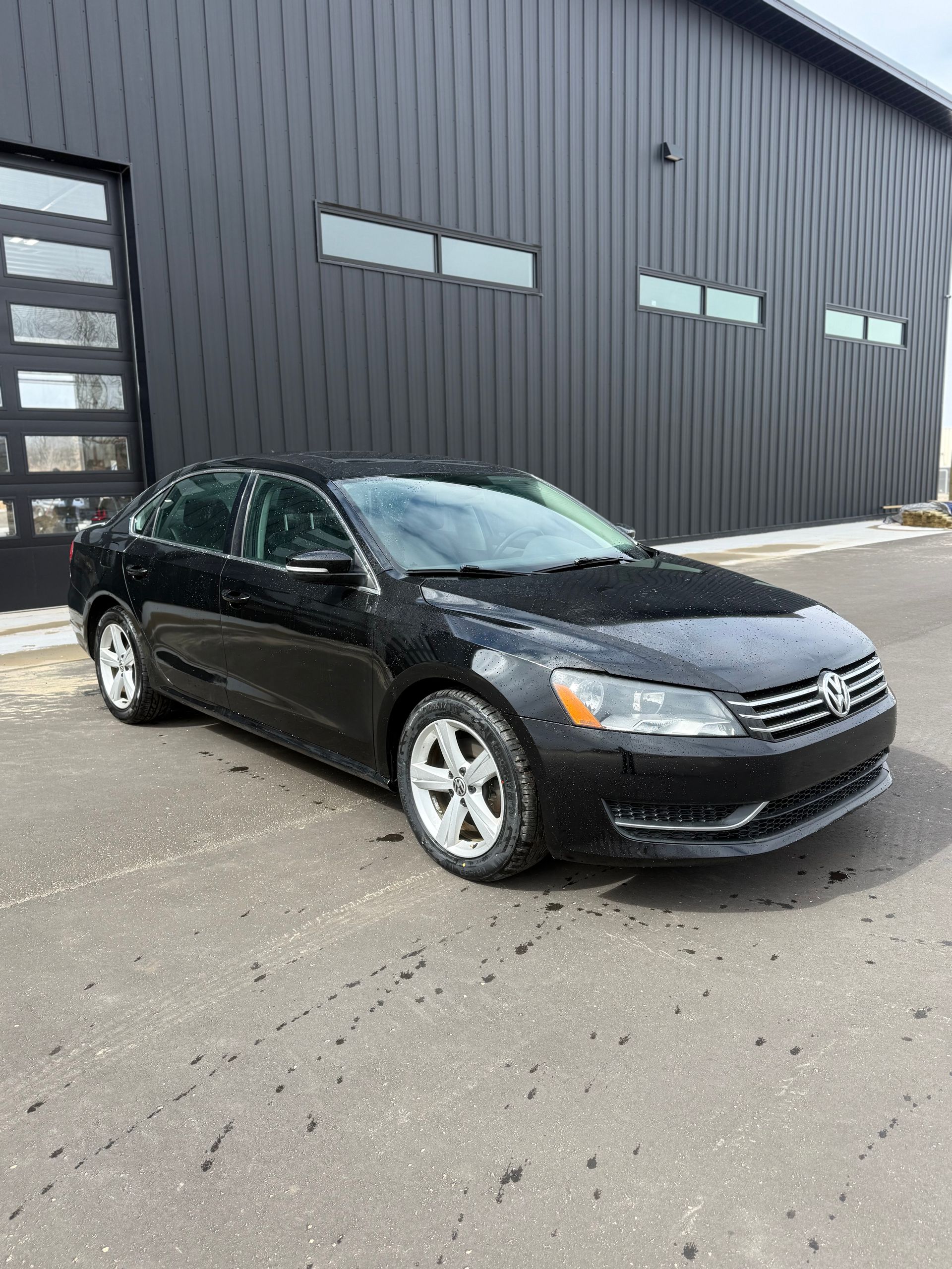 A black Volkswagen Passat sedan parked on a grey asphalt lot in front of a dark metal building with glass garage doors.