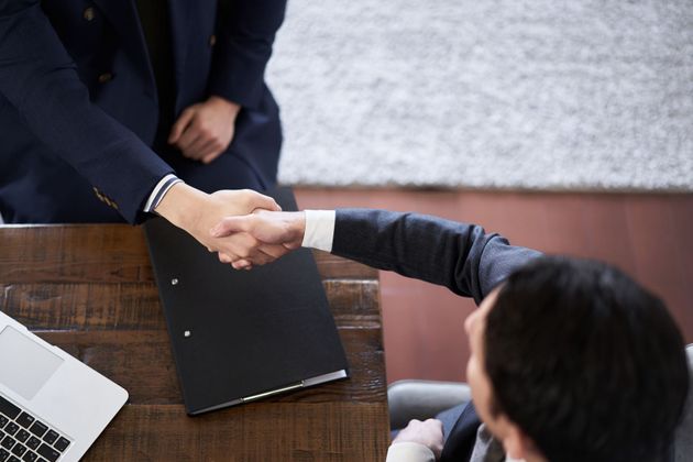 Two men in suits shaking hands over a desk with a laptop and folder.