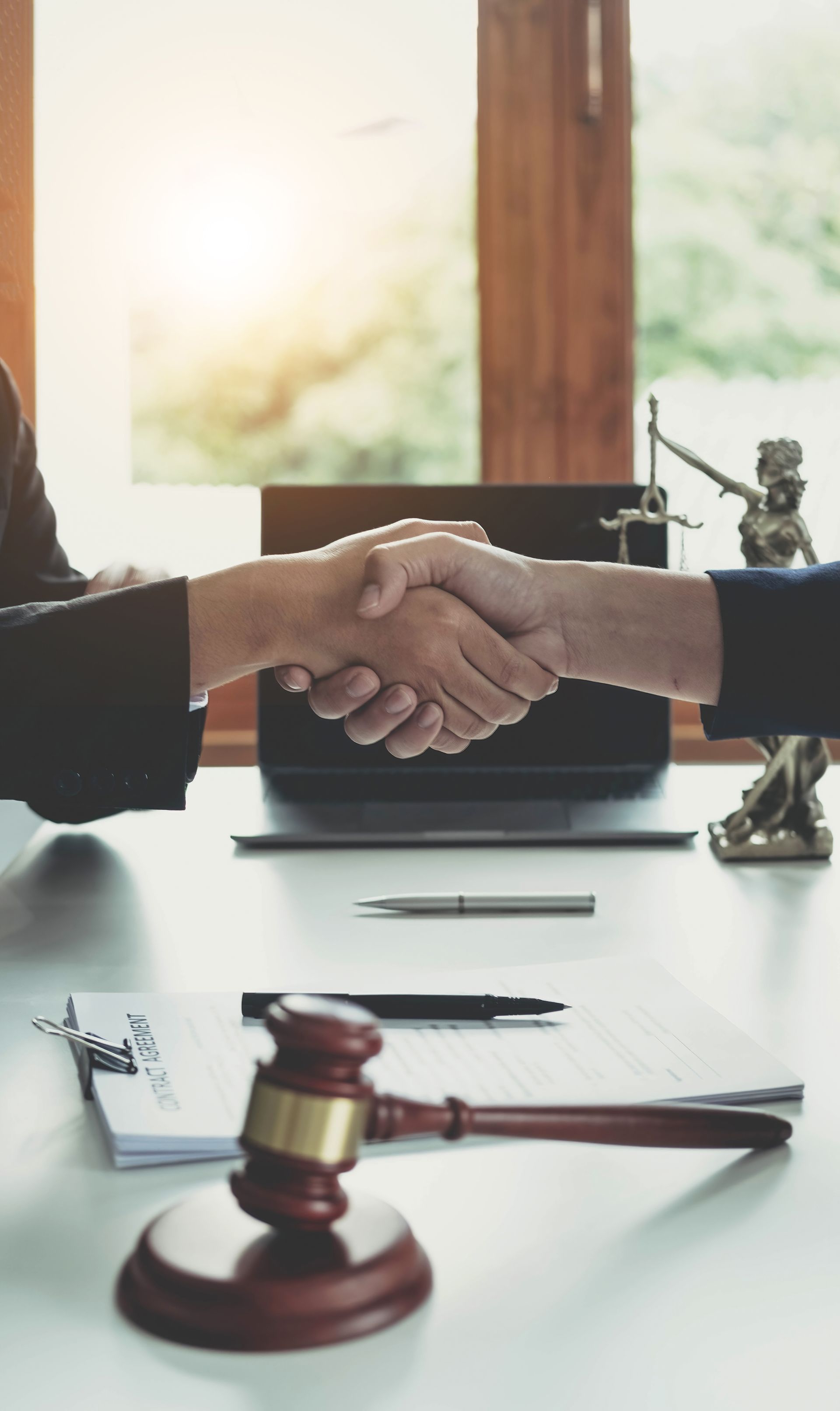 Two people shake hands over a desk, with a gavel and scales of justice in the foreground.