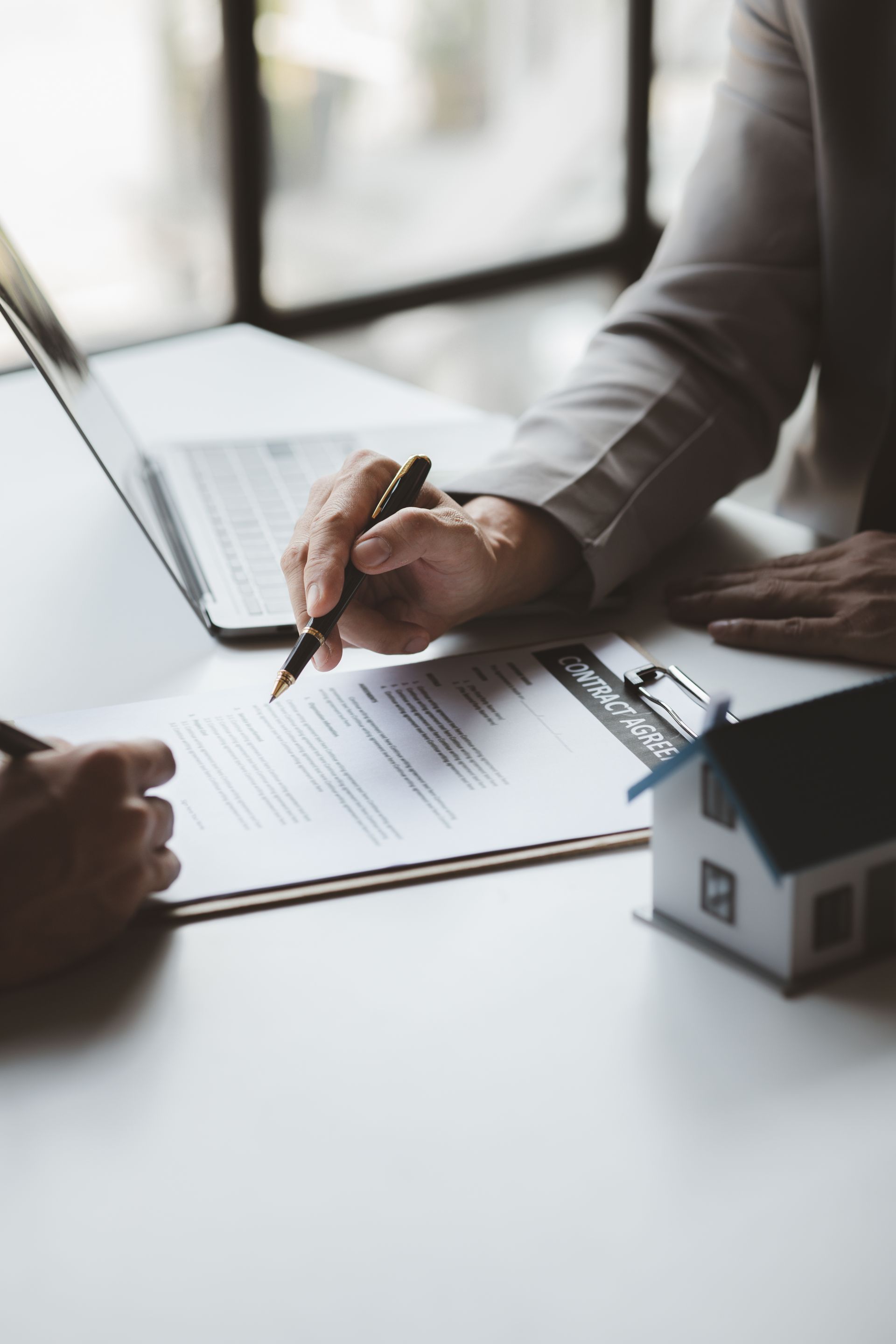 Person signing real estate contract with a pen, miniature house on table, laptop nearby.