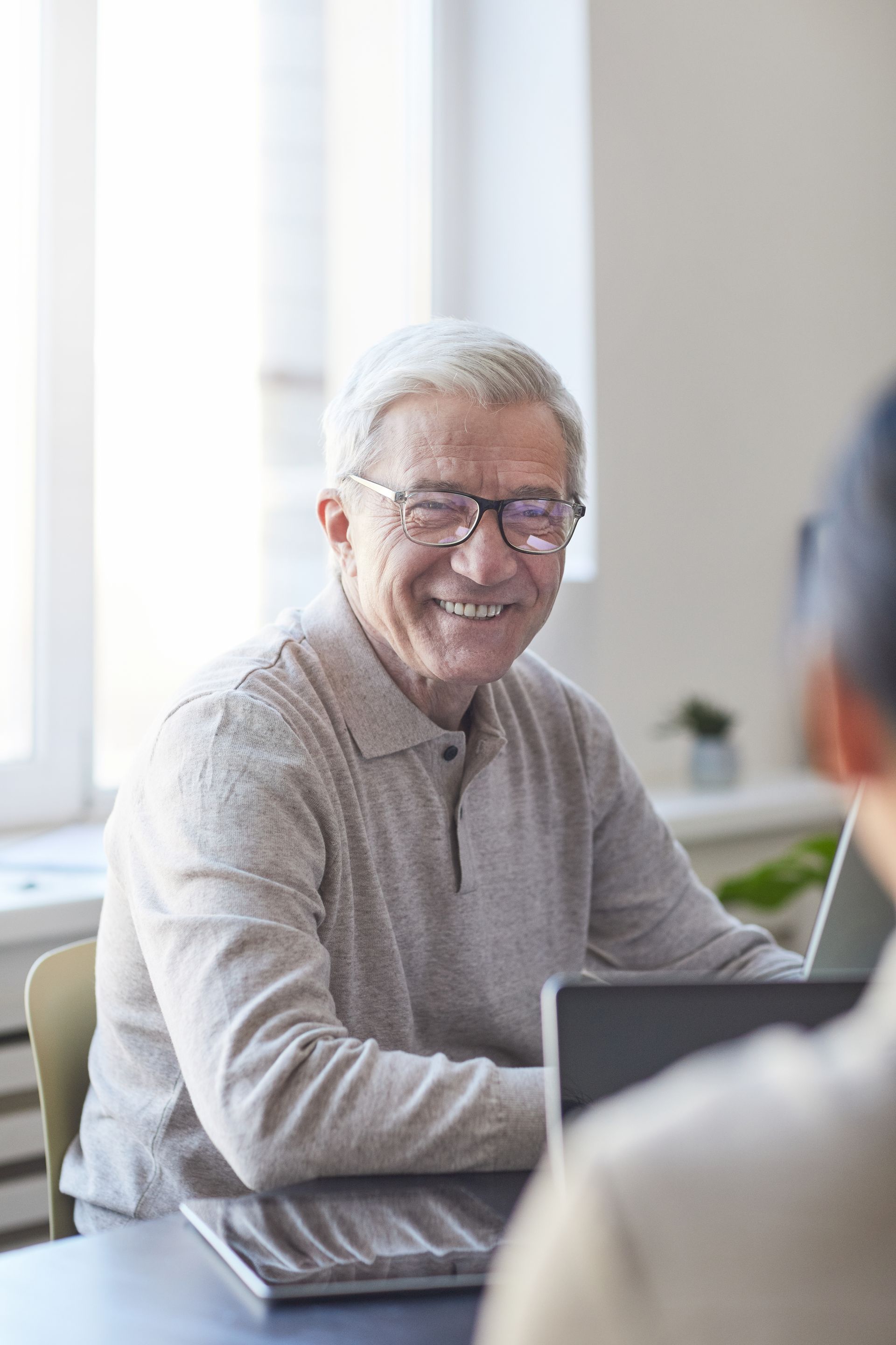 Smiling elderly man with glasses at a table, working on a laptop.