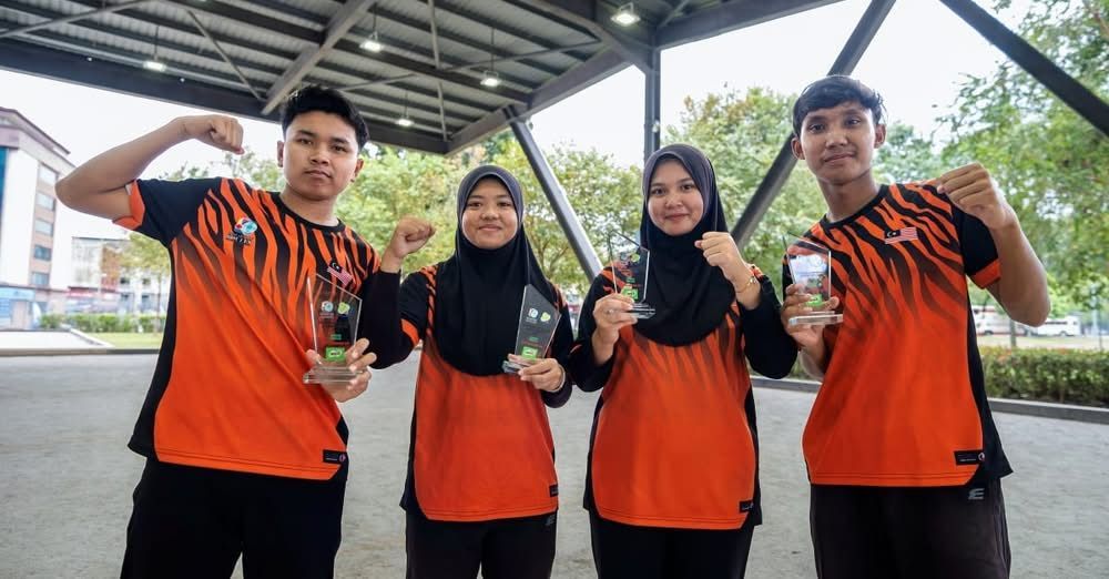 Man in orange shirt throws a metal ball during a pétanque game. Malaysia vs. Thailand scoreboard visible.