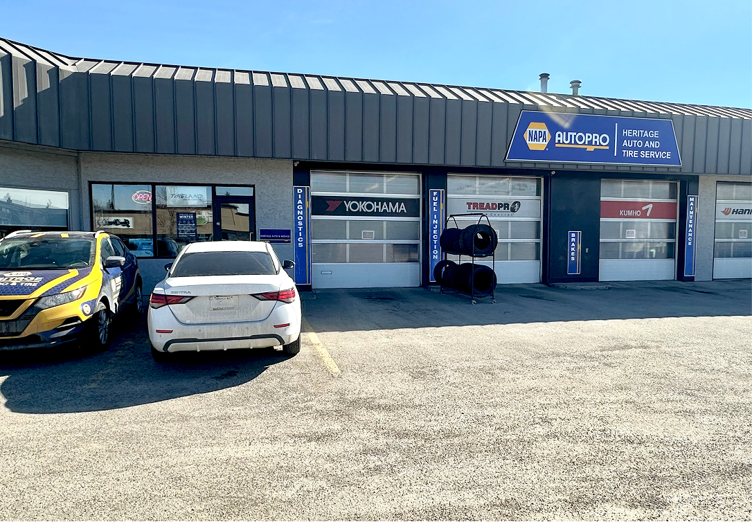 A commercial auto shop exterior with two cars parked in front of three service bay doors under a sunny sky. | Heritage Auto & Tire Service