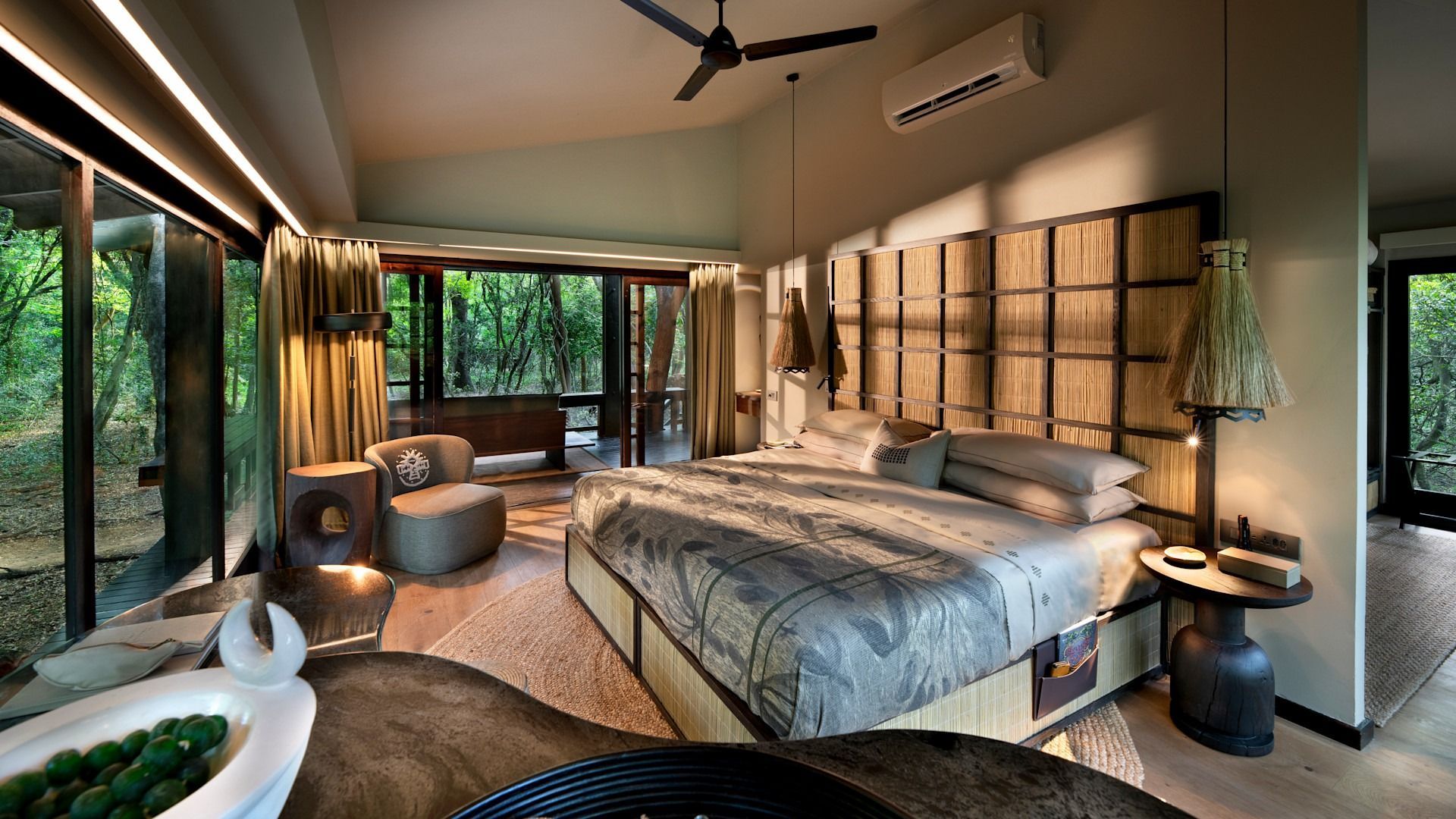 Luxurious bedroom with king bed, open to a jungle view. Neutral tones, woven headboard, and decorative lighting.