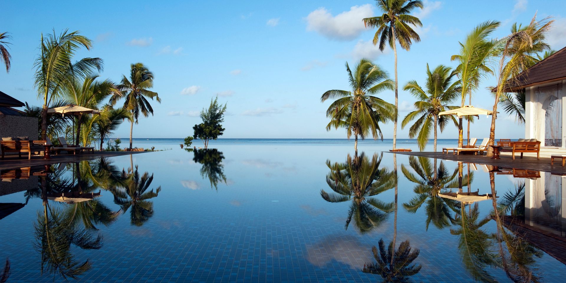 An infinity pool reflects palm trees and a clear blue sky, overlooking a calm ocean at a tropical resort. The Residence Zanzibar
