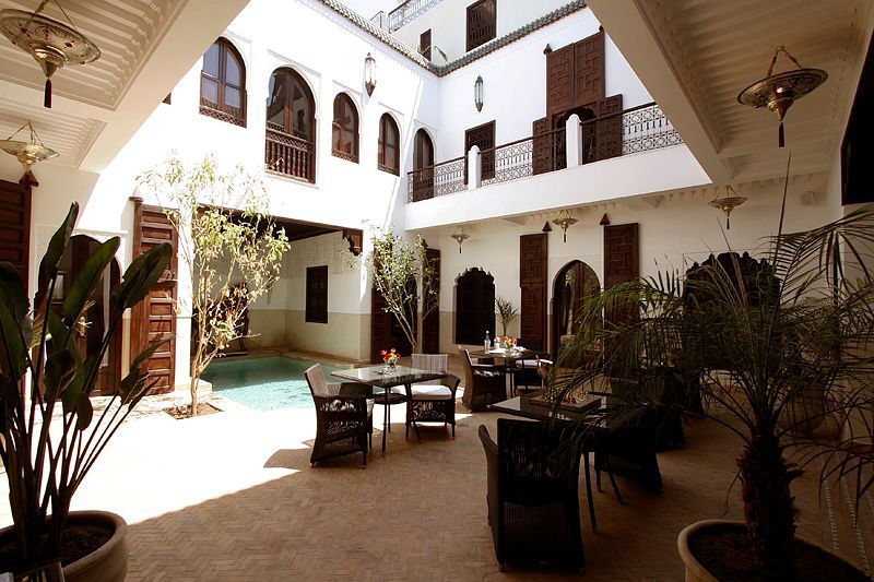 A sunlit courtyard in a Moroccan-style riad featuring a small pool, patio furniture, potted plants, and arched doorways.
