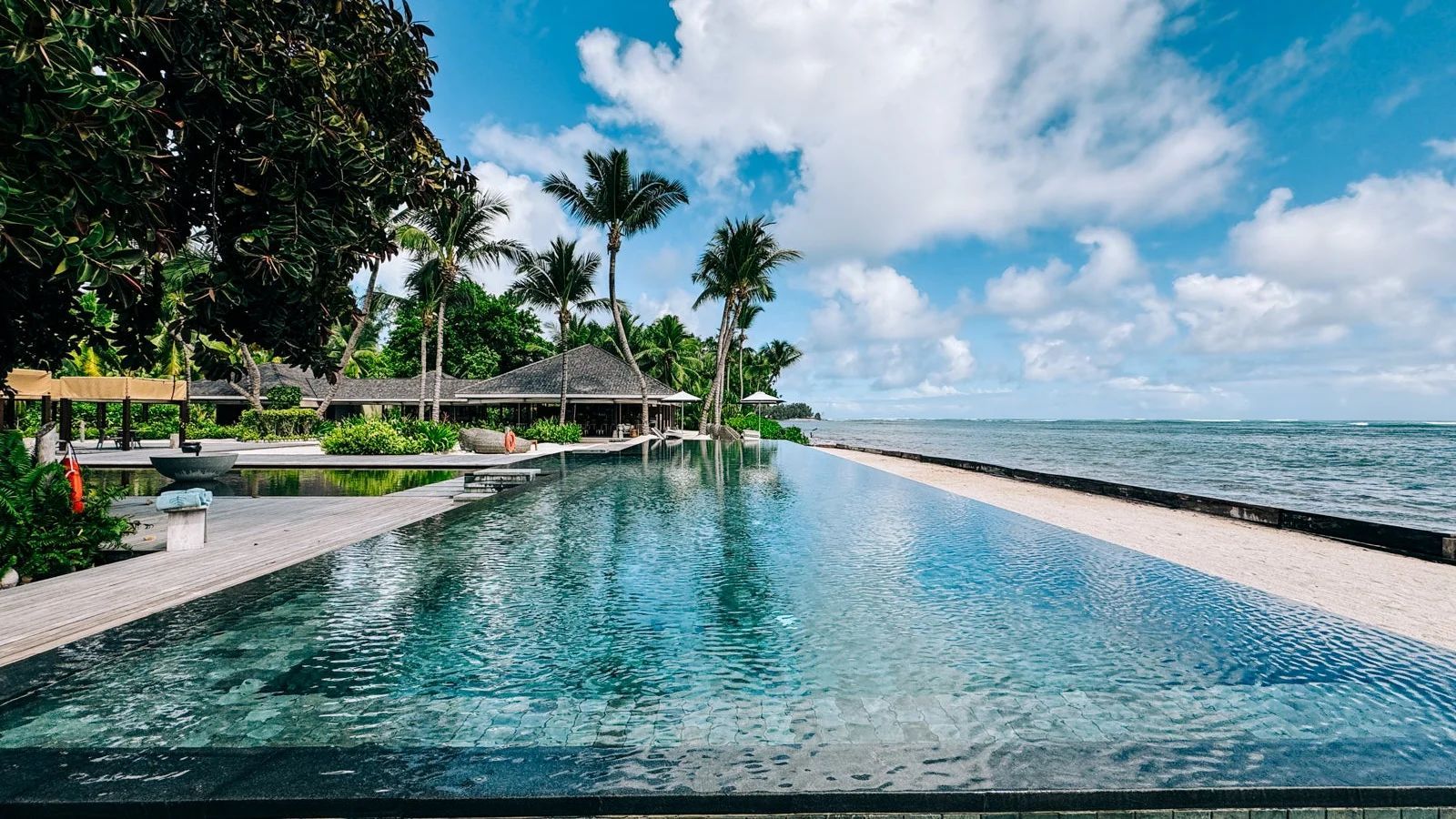 An infinity pool overlooking the ocean, framed by tropical palm trees and lush greenery under a bright, cloudy blue sky. Four Seasons Resort Seychelles at Desroches Island