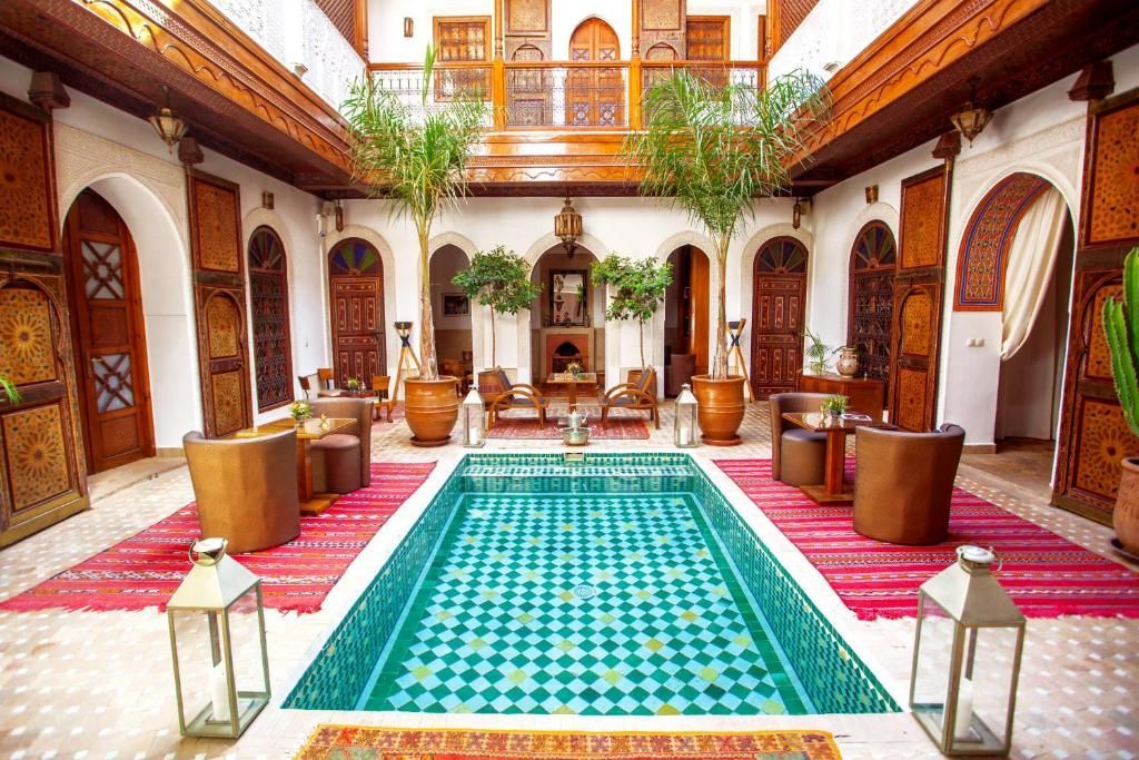 A central turquoise tiled swimming pool in a Moroccan-style courtyard with wooden doors, plush chairs, and pink rugs.