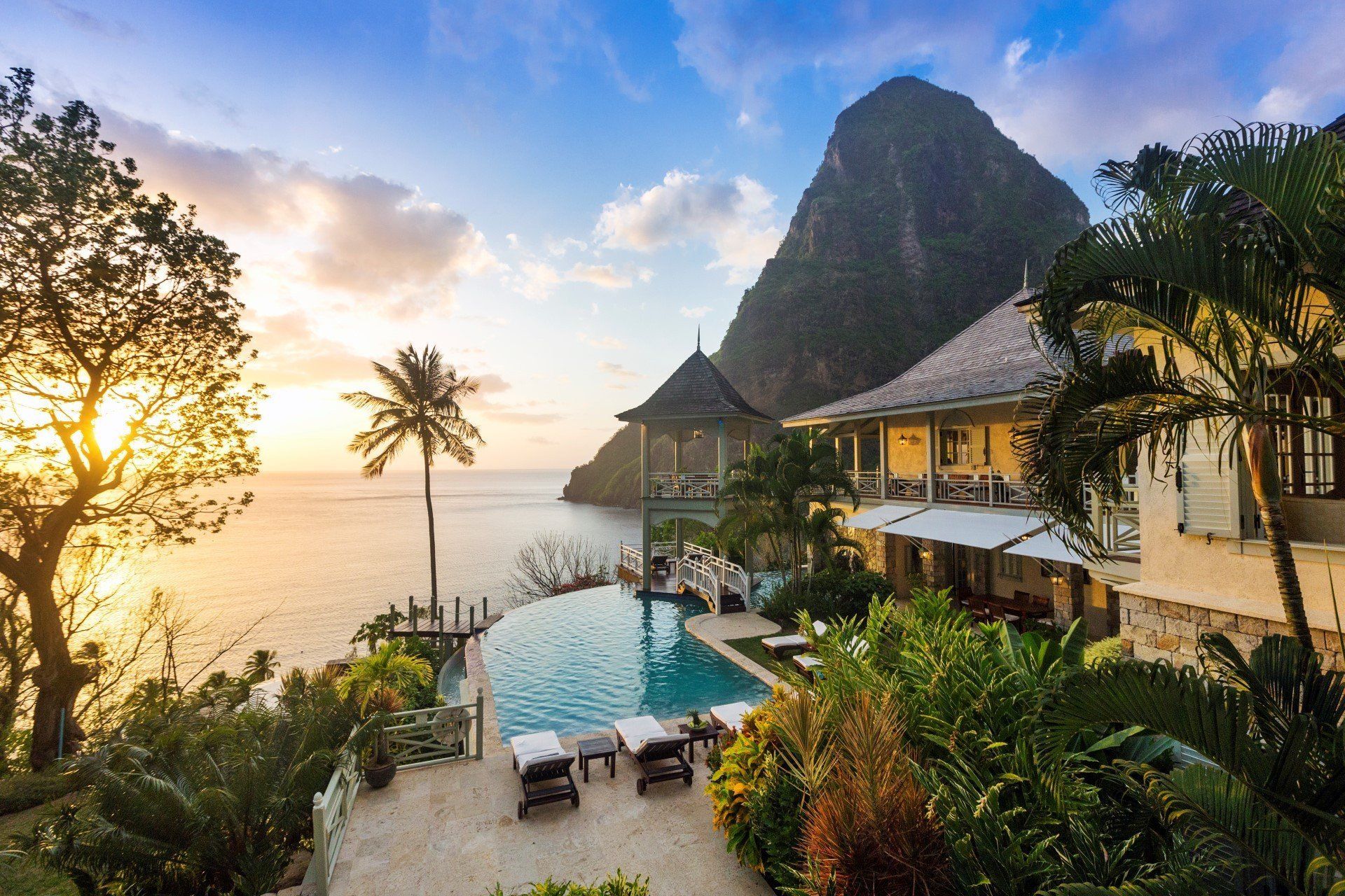 Infinity pool overlooking ocean, with Pitons mountains in the background at sunset. Lush greenery surrounds resort buildings. 4. Arc En Ciel Villa – St Lucia