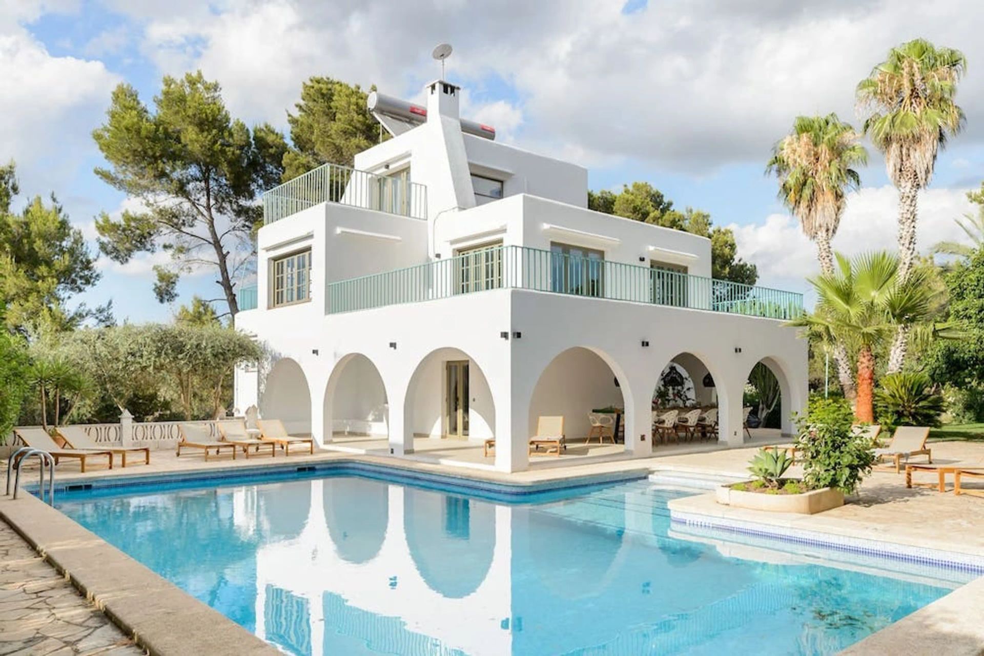 A white, multi-story Mediterranean villa with arched porches overlooking a bright blue swimming pool in a sunny garden.