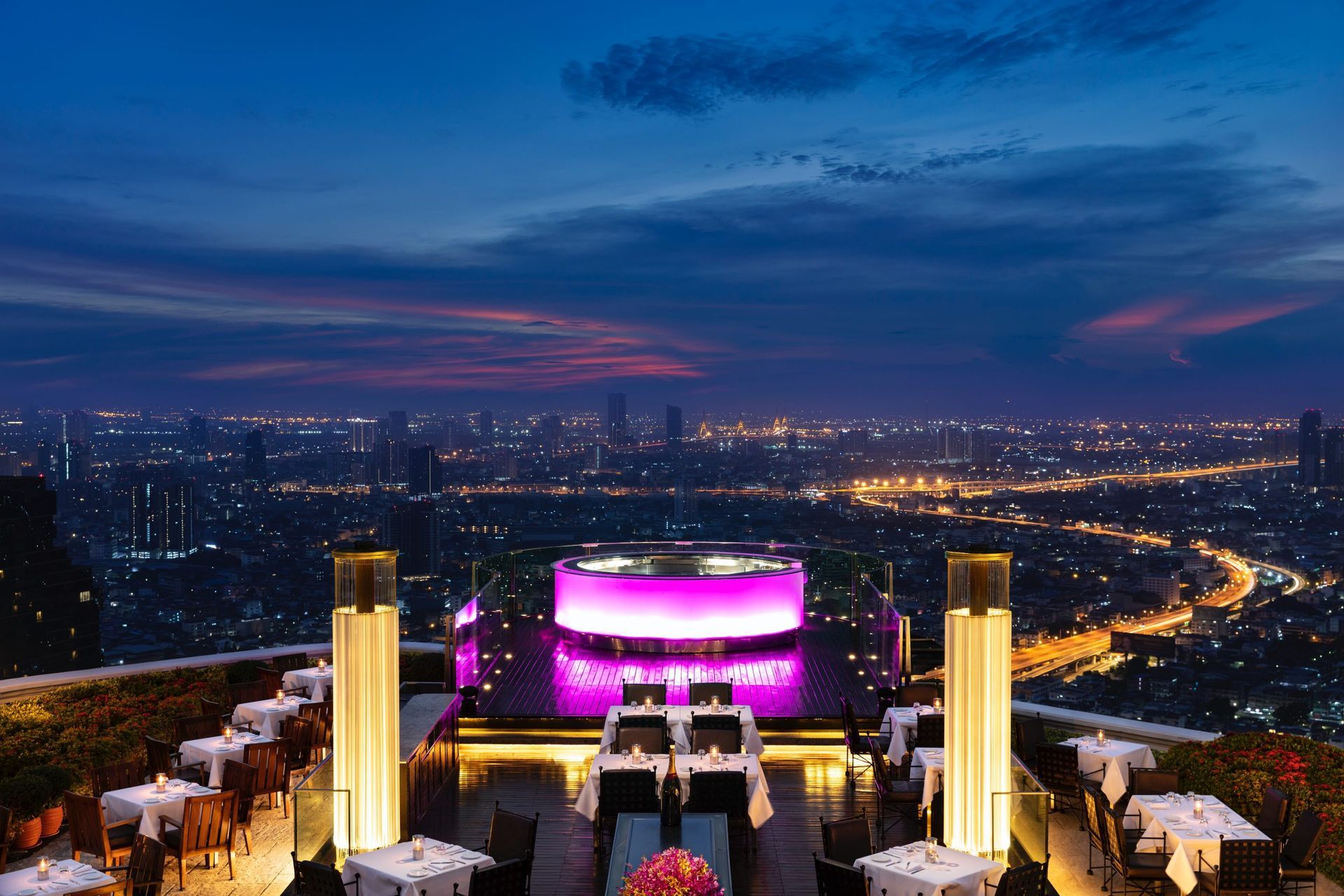 Rooftop restaurant at twilight with city skyline views, tables set, illuminated bar, and vibrant purple and gold lighting.