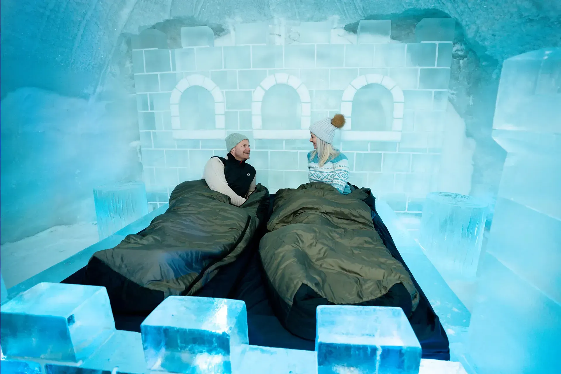 Two people sitting up in sleeping bags on a bed inside a room constructed entirely from blocks of blue ice. Snowhotel Kirkenes, Kirkenes
