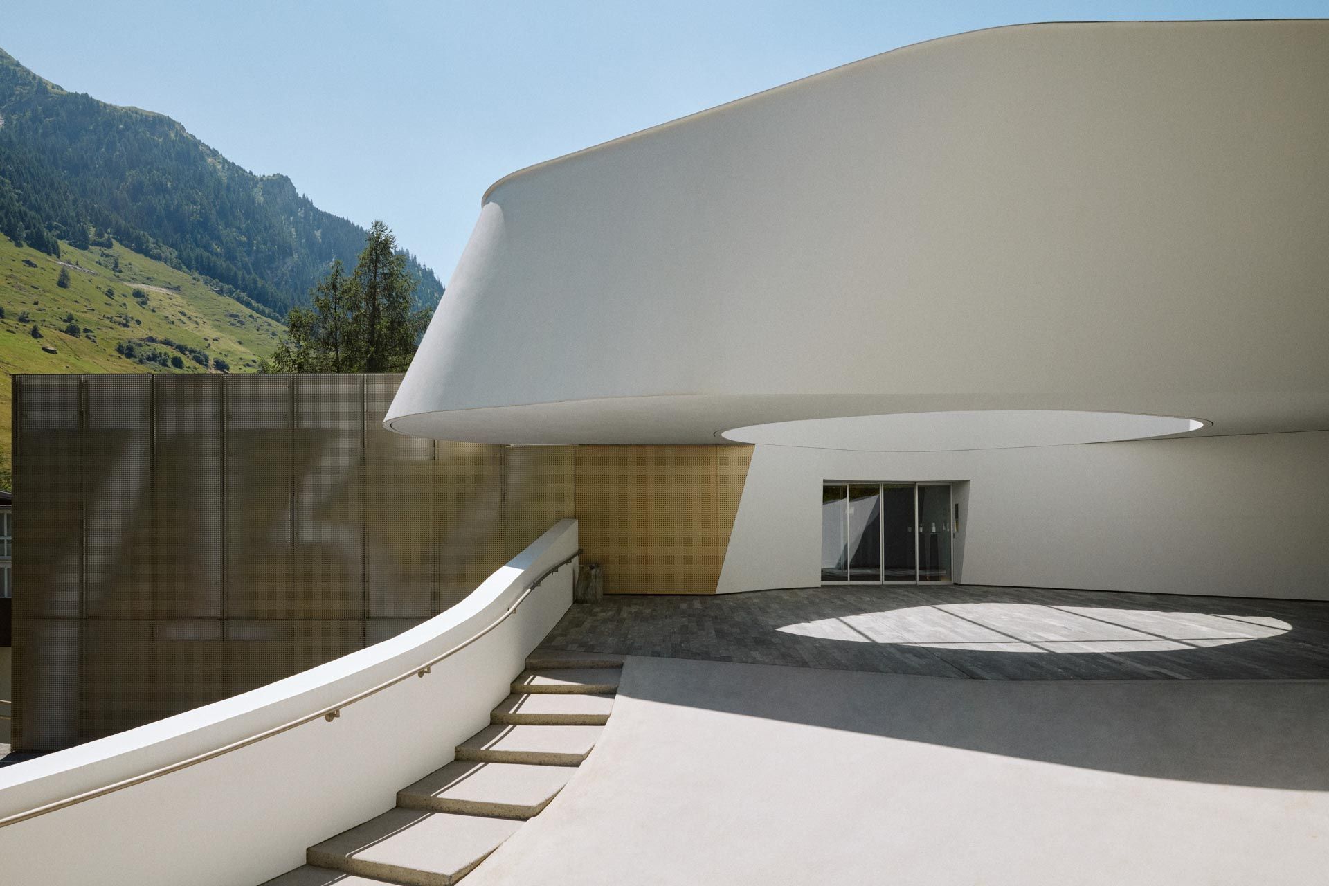 Modern white building with curved facade and entryway, steps, and mountain backdrop. 7132 Hotel & Therme Vals (Vals)