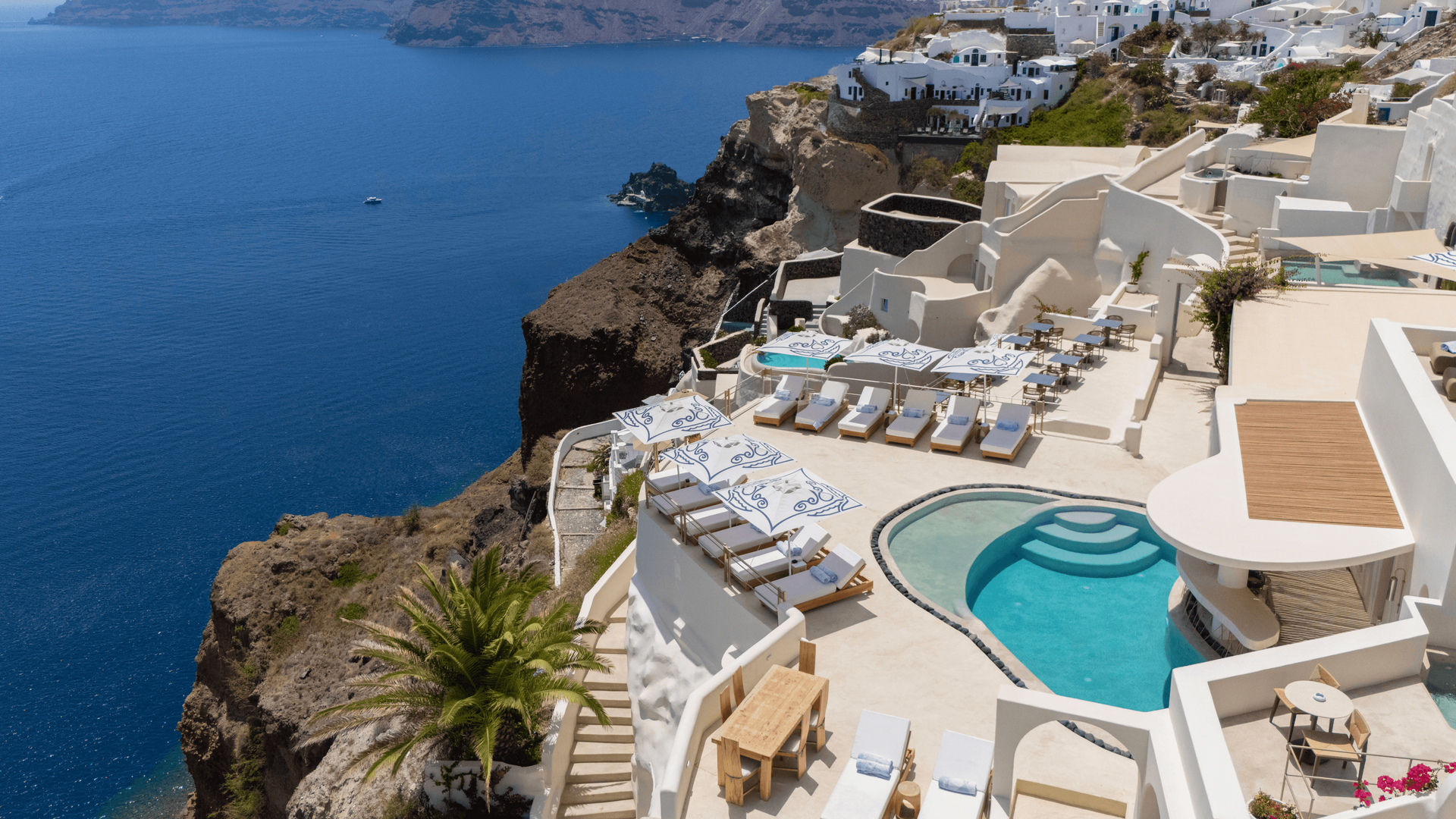 A high-angle view of a white, terraced luxury hotel on a rocky cliff overlooking the deep blue sea in Santorini, Greece. Santo Pure Oia Suites & Villas