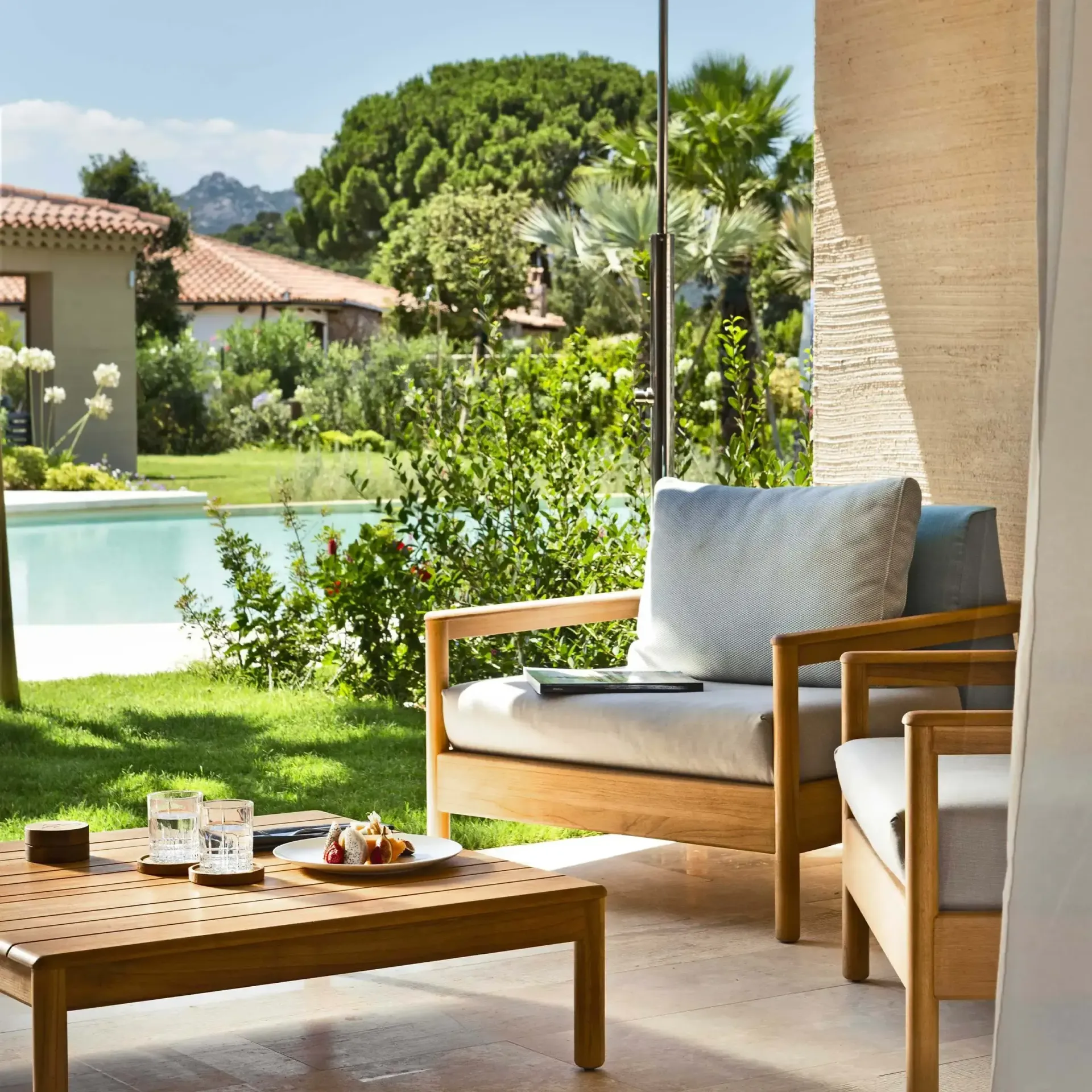 Patio seating beside a pool with a lawn, coffee table, and sunlit garden view 7Pines Resort Sardinia
