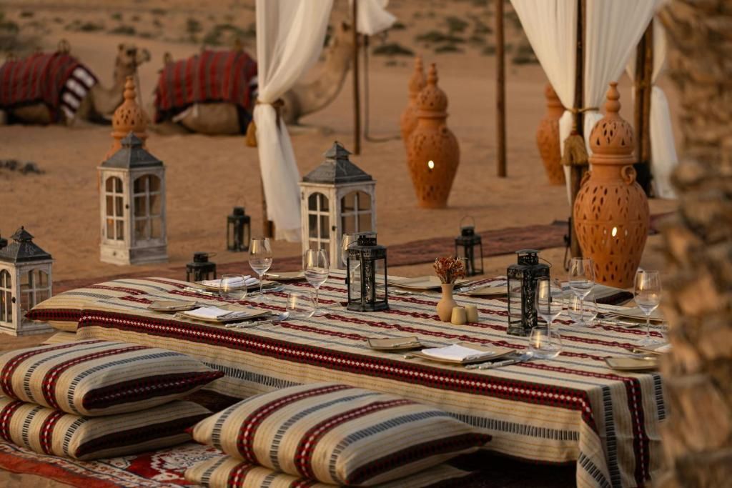 Desert dining setup with lanterns, cushions, and pottery. Camels in background.
