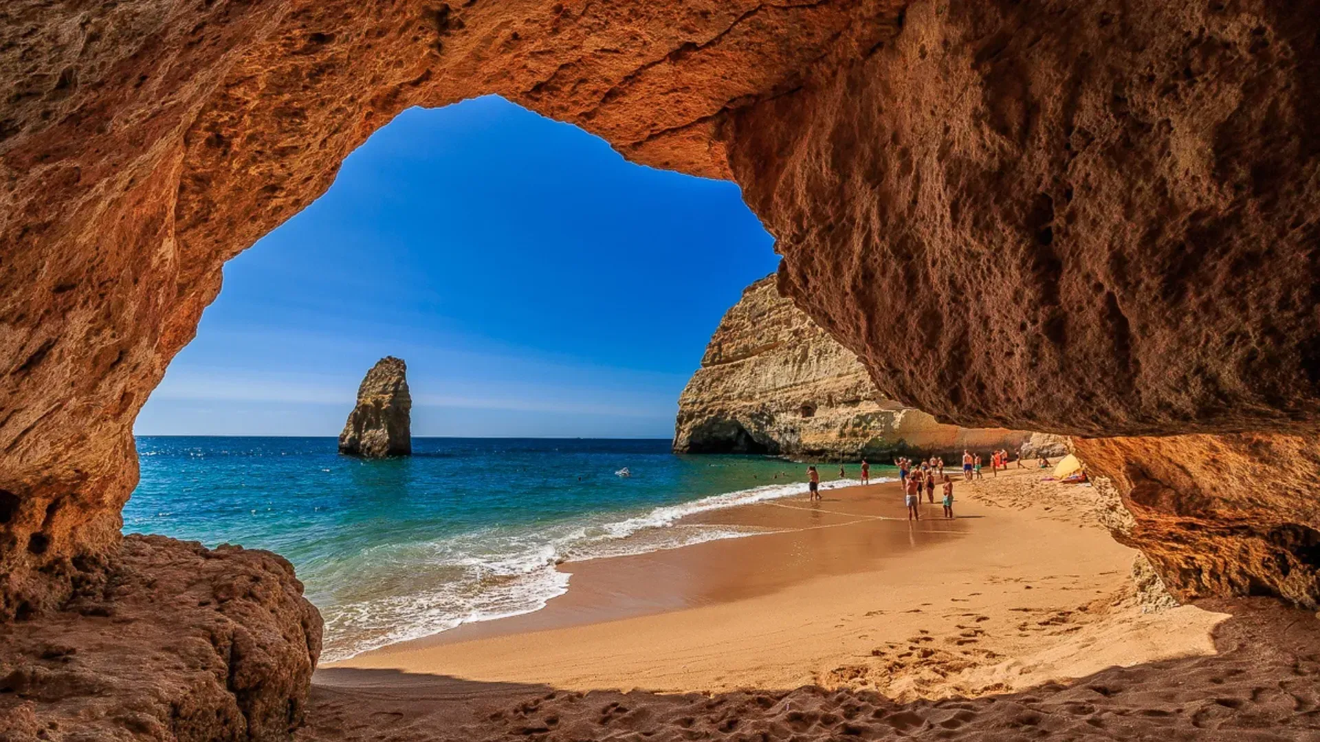 Beach scene viewed through a rock arch: turquoise water, sandy beach, people, cliffs, and a sunny blue sky. Algarve Family Holiday