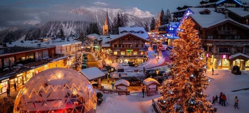 A snow-covered town at dusk, with buildings, a Christmas tree, and mountains in the background.