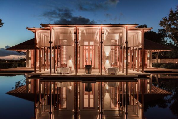 Elegant, illuminated building reflecting in a still pond at dusk. Tables set inside, warm lighting, serene setting. Amanyara, Turks and Caicos