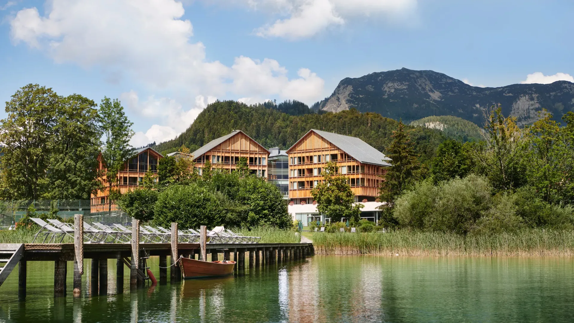 Wooden buildings on a lake with a dock and boat, mountain in the background. Sunny day.