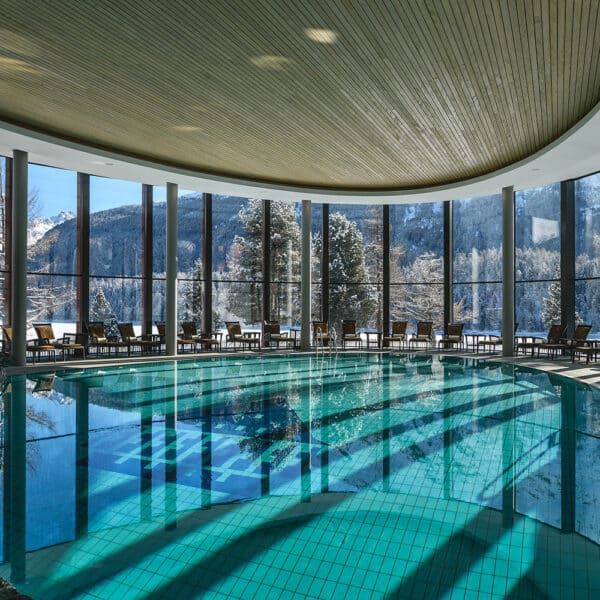 Indoor pool with large windows, snow-covered trees outside. Blue water reflects light. At Badrutt’s Palace Hotel