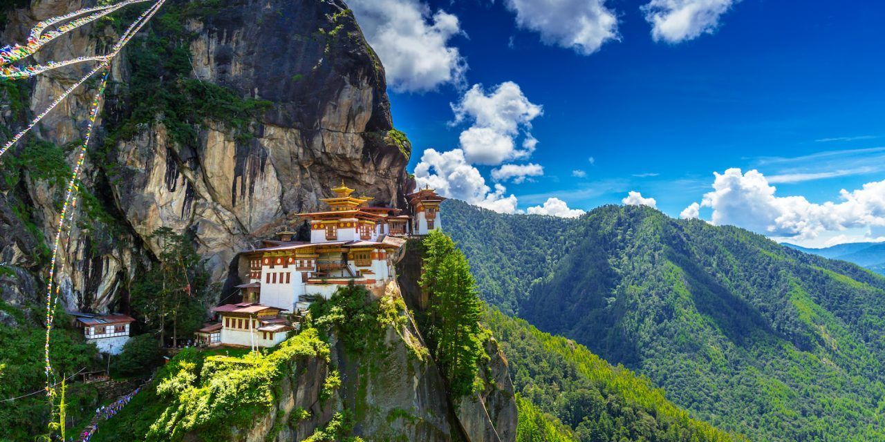 Taktshang Monastery clings to a cliff face in Bhutan. Lush green mountains and a blue sky with clouds frame the scene.