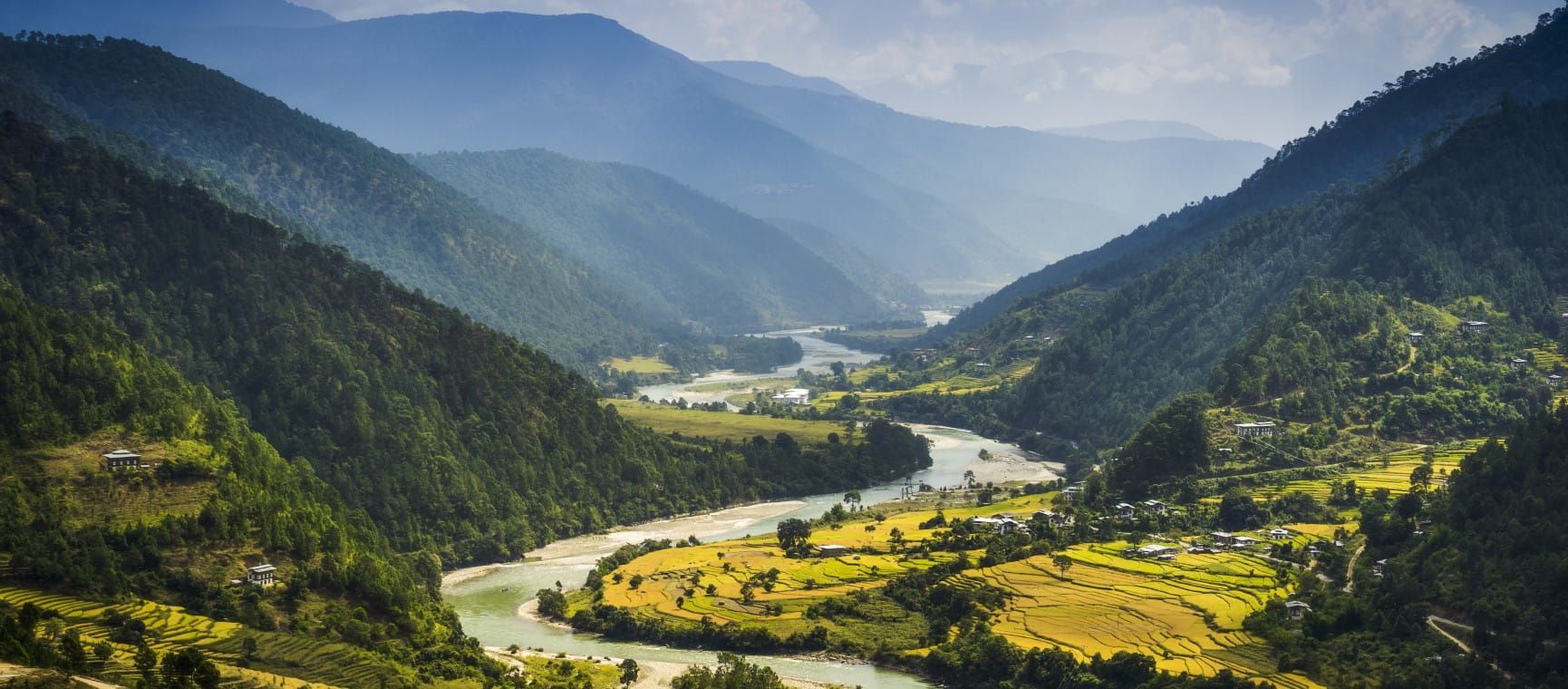 River winds through a green valley framed by forested mountains under a hazy sky. Bhutan
