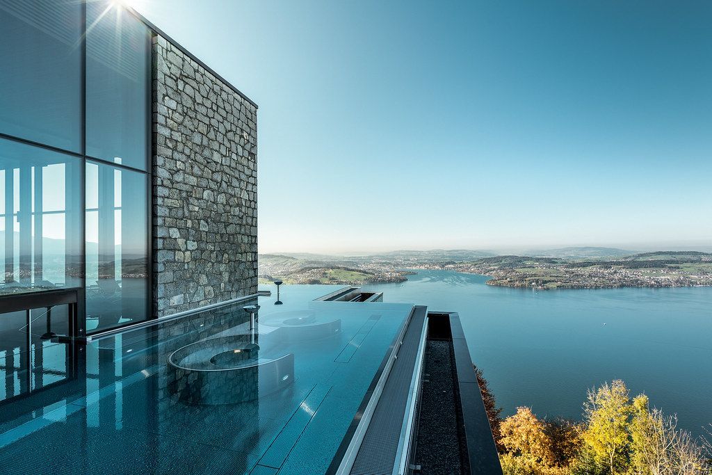 Infinity pool overlooking a lake and distant cityscape; modern building with stone and glass facade; sunny day. Bürgenstock Resort