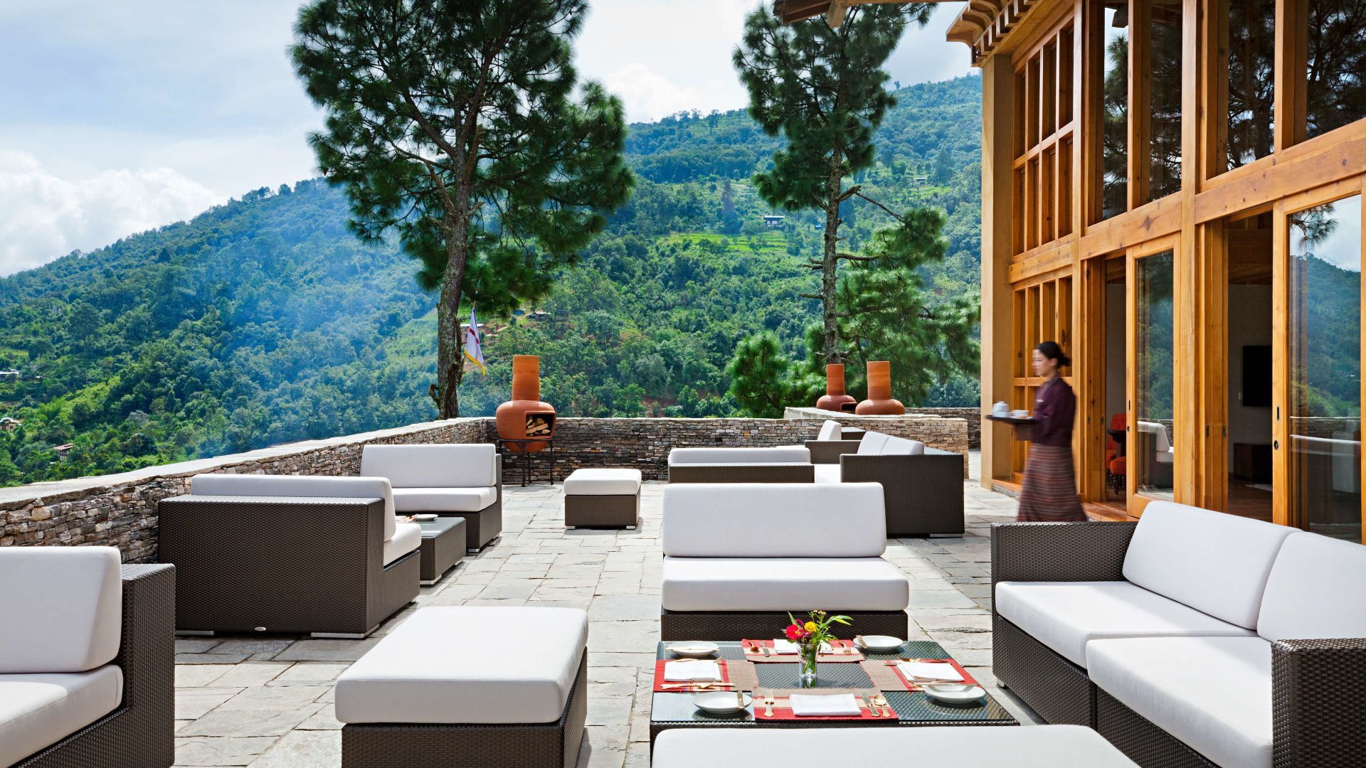 Outdoor lounge area with white sofas, overlooking a green mountain range; a person carries a tray. COMO Uma Punakha