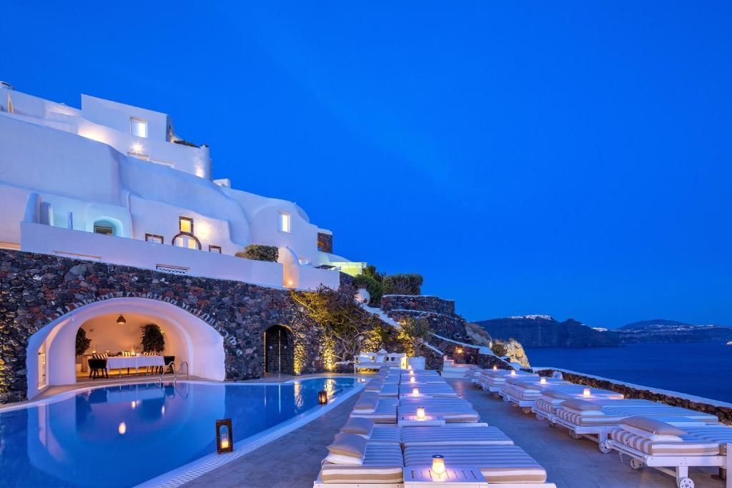 Whitewashed buildings with a pool and lounge chairs overlooking the sea at dusk in Santorini, Greece. Canaves Oia Suites