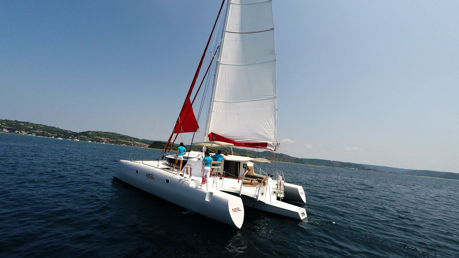 A white catamaran sailing on blue water with a red sail, crew on deck, under a clear sky.