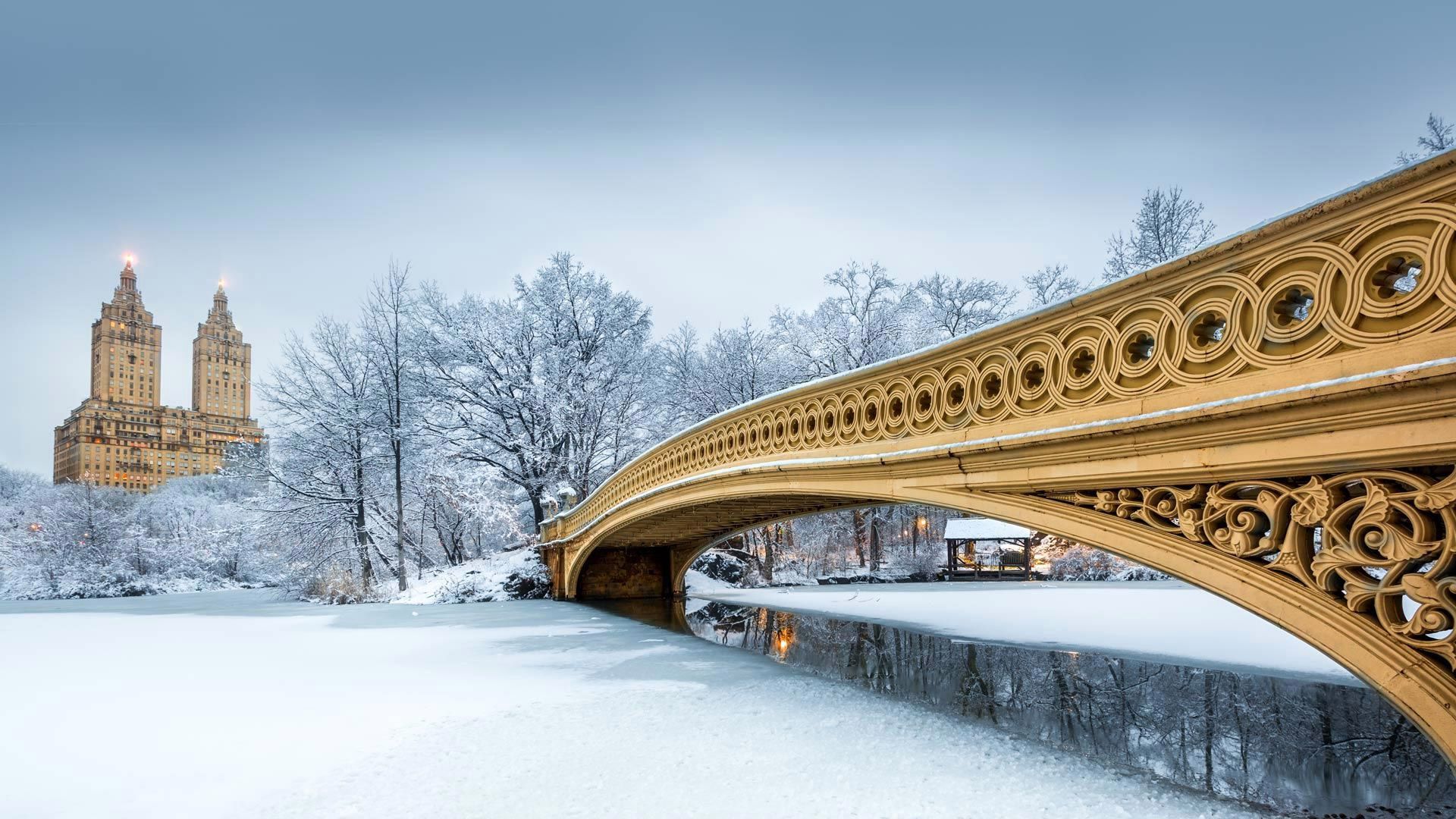 Snowy Central Park with Bow Bridge, a yellow ornate bridge, and distant twin-towered building. Central Park