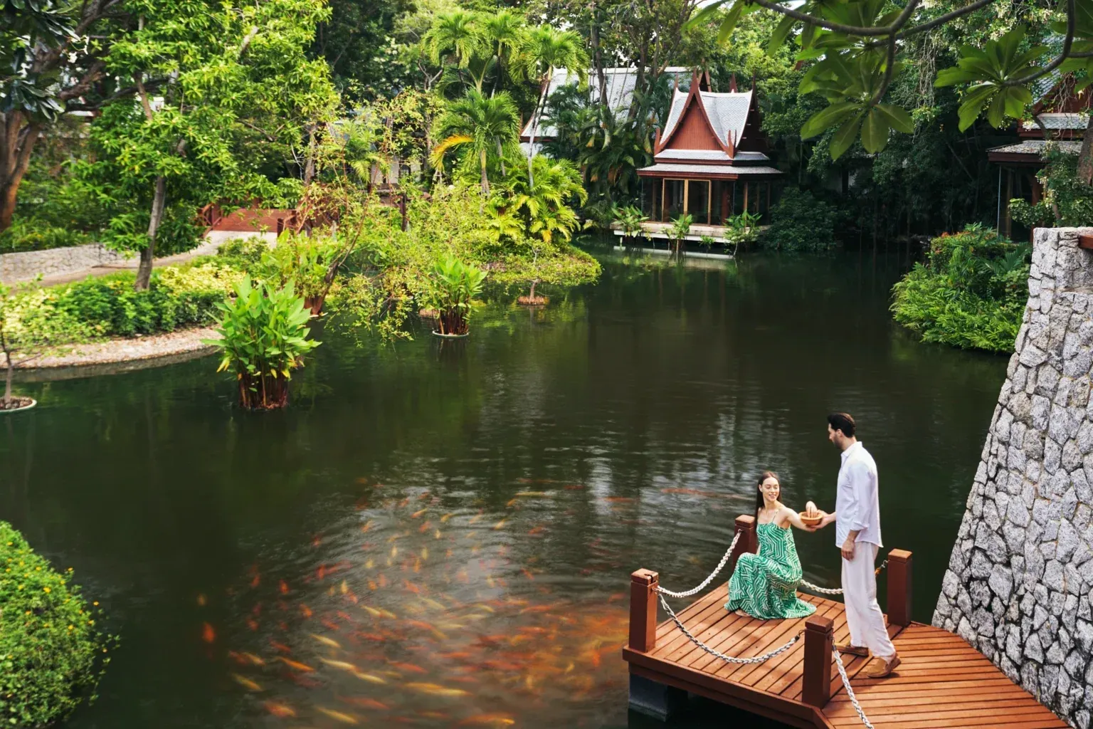 Chiva-Som, Hua Hin A couple stands on a wooden dock over a pond with many koi fish, surrounded by lush greenery and buildings in a tropical setting.