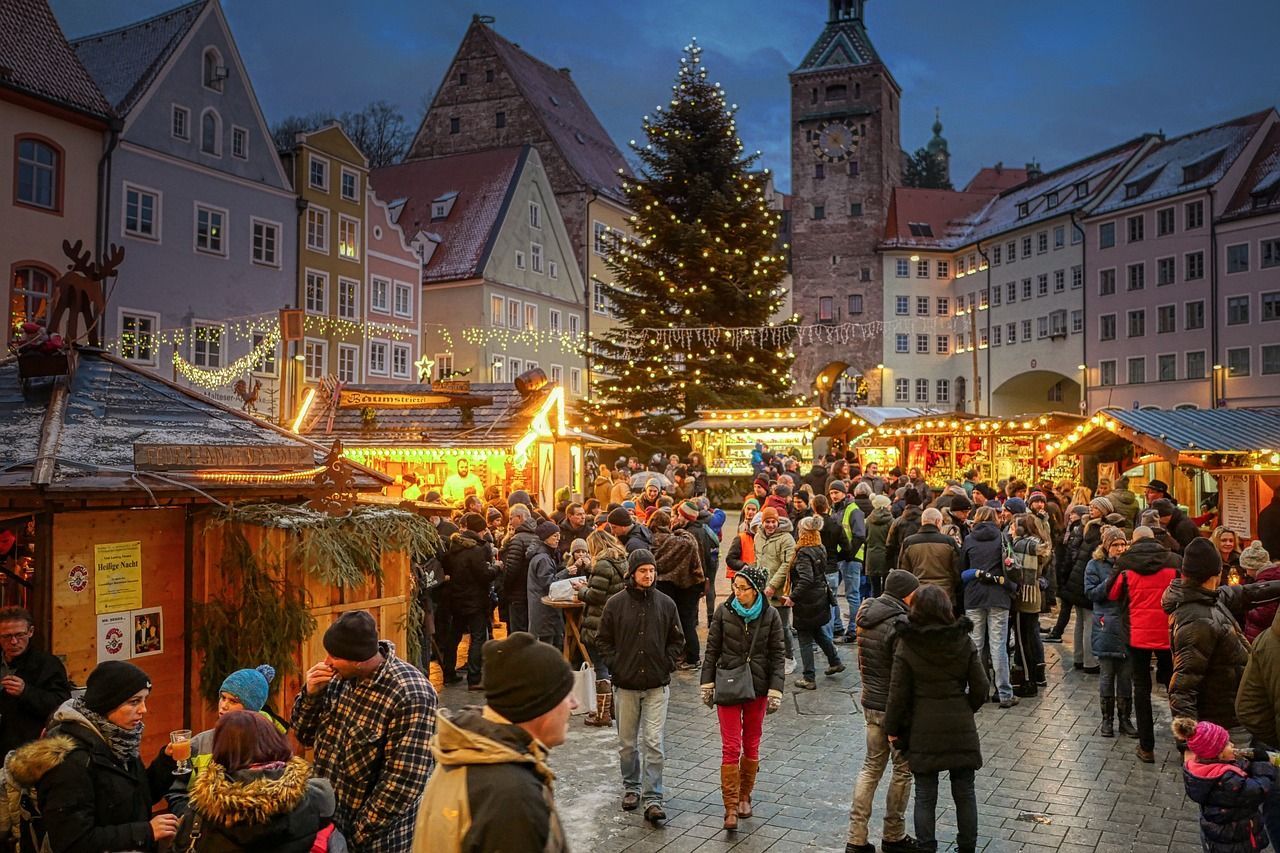 Christmas market in a European square at dusk, with crowds, stalls, a lit Christmas tree, and historic buildings.