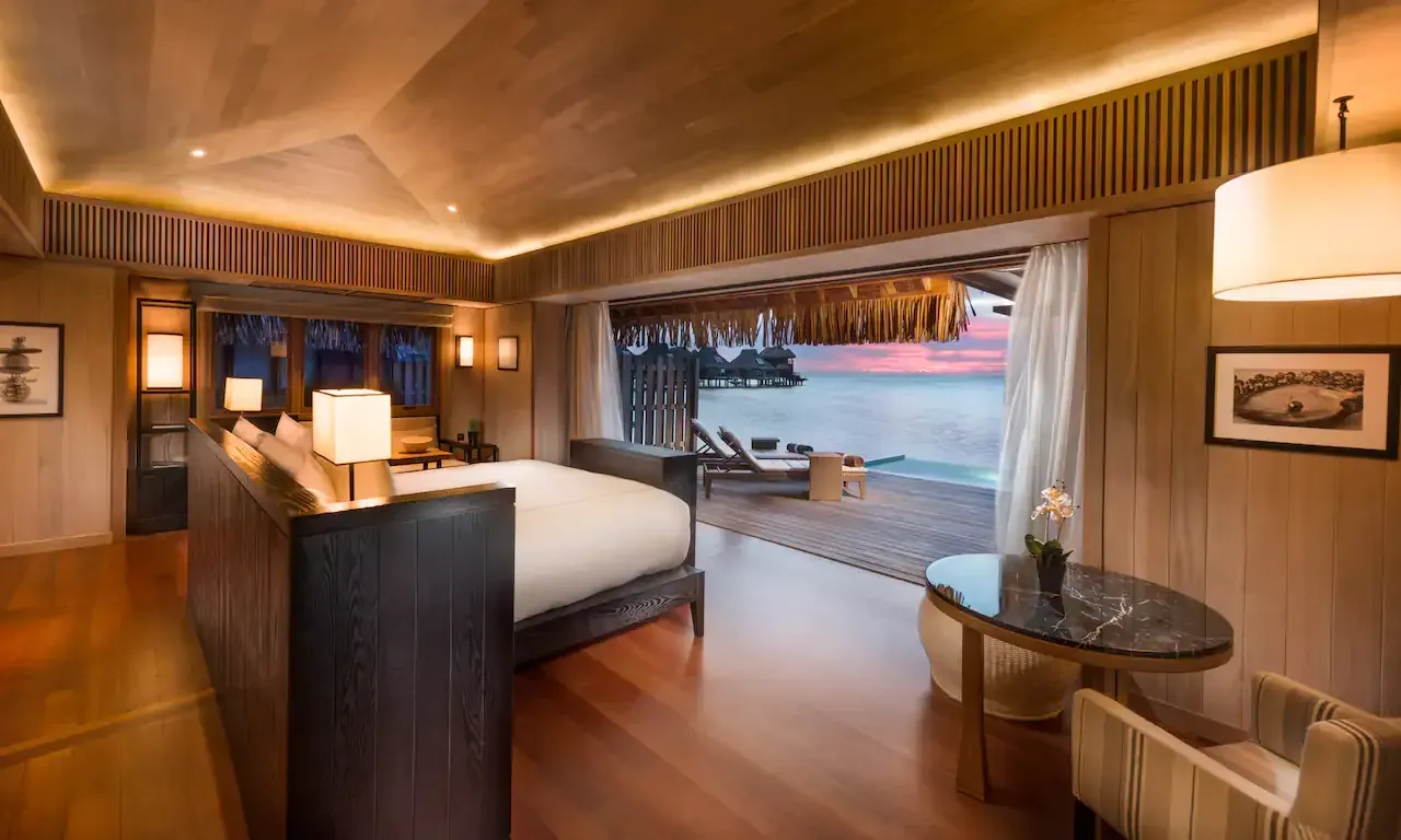 Wooden luxury hotel room overlooking the ocean at sunset. Features a bed, table, and access to a private deck. Conrad Bora Bora Nui