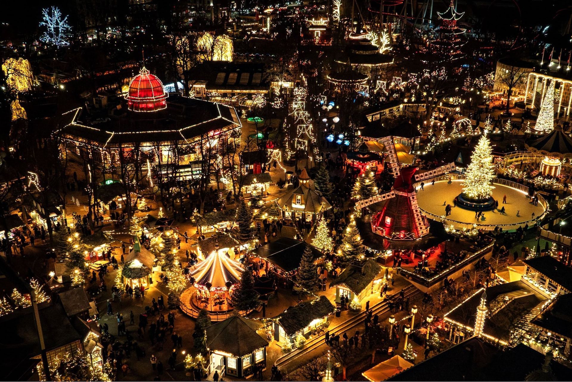 Overhead night view of a brightly lit Christmas market with rides, stalls, and a large illuminated tree. Copenhagen