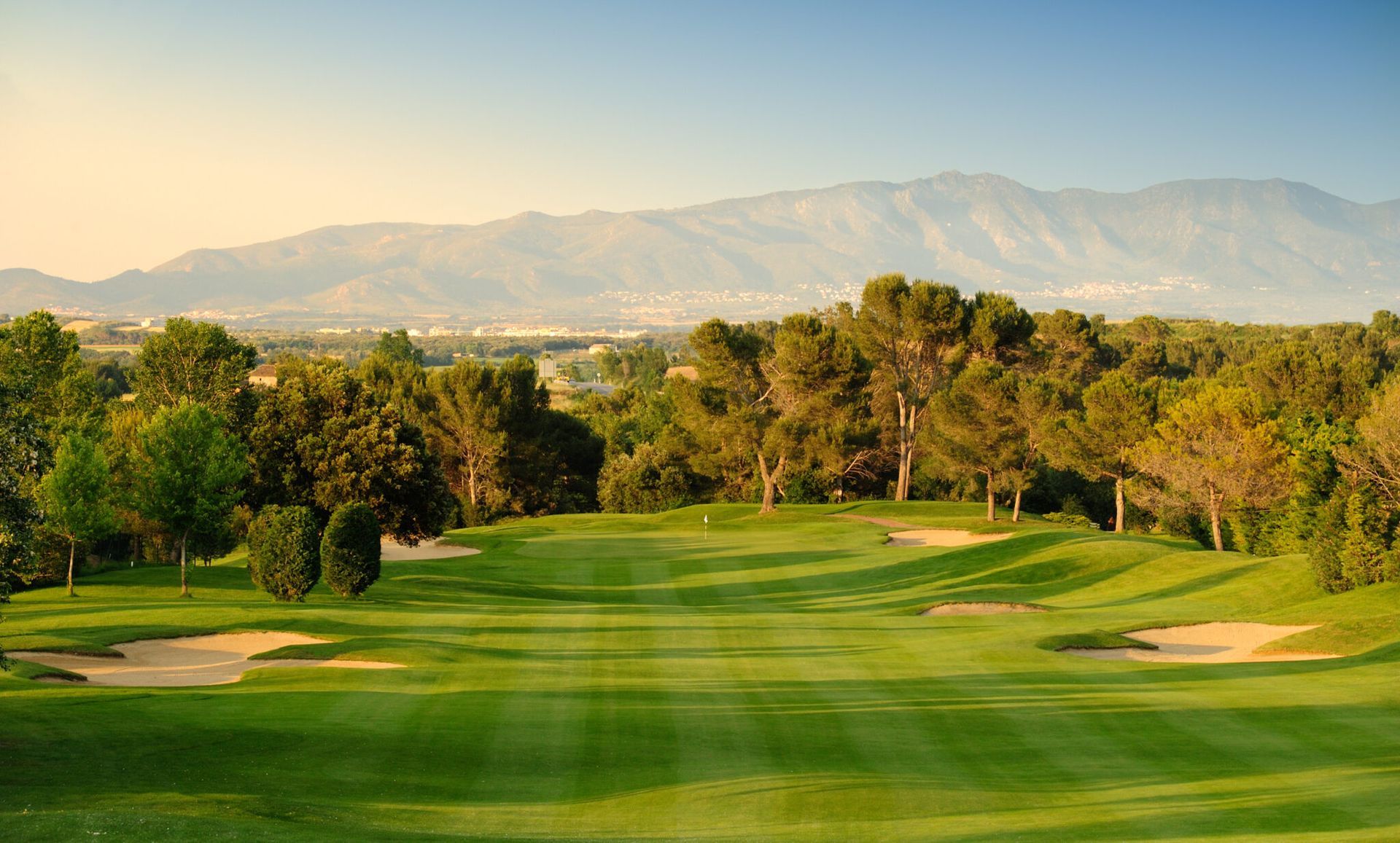Green golf course with trees, bunkers, and mountains in the background under a blue sky. Costa Brava