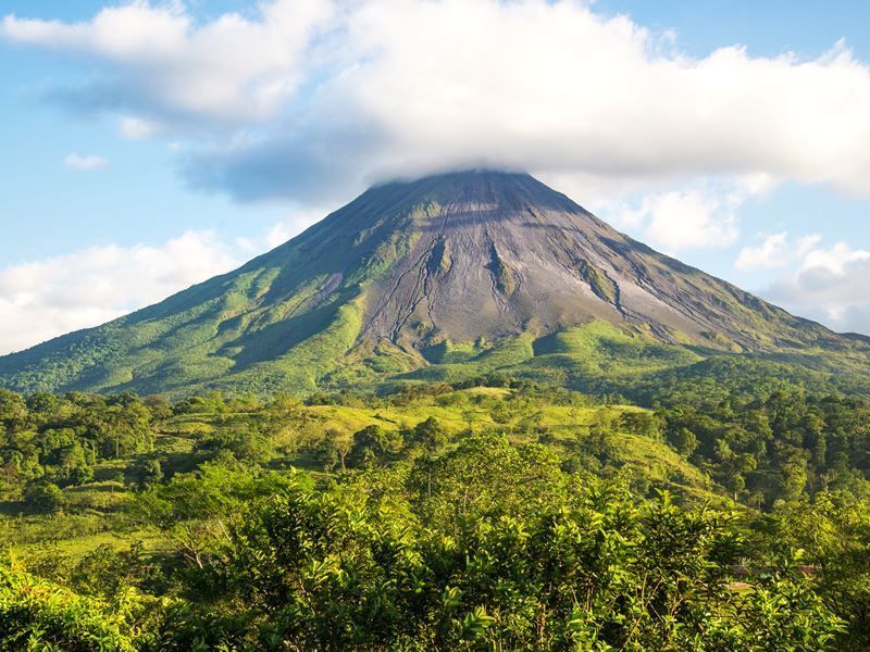 A conical volcano rises above lush green vegetation, partly obscured by a cloud.