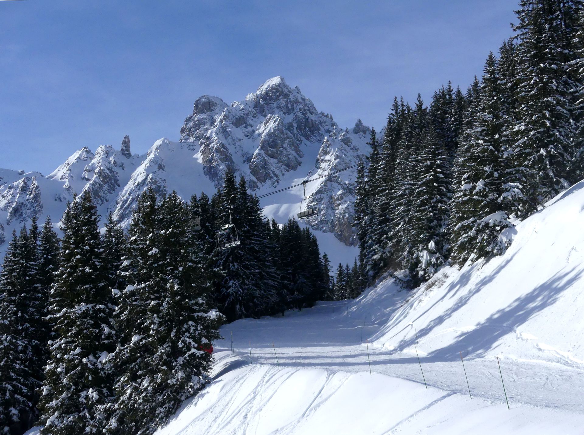 Snow-covered evergreen trees border a turquoise lake, with a wooden pier extending into the water under a clear blue sky. Courchevel