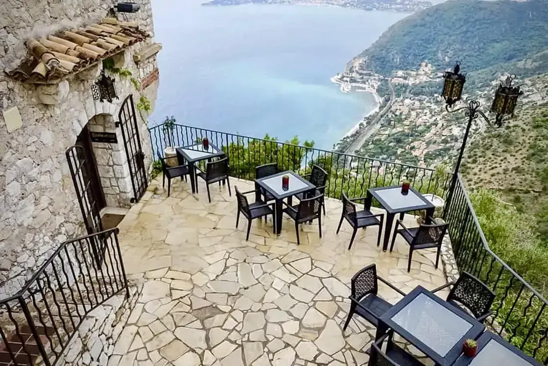 Restaurant patio overlooking the sea. Stone walls and floor, black tables and chairs. Scenic view. Èze & Grasse