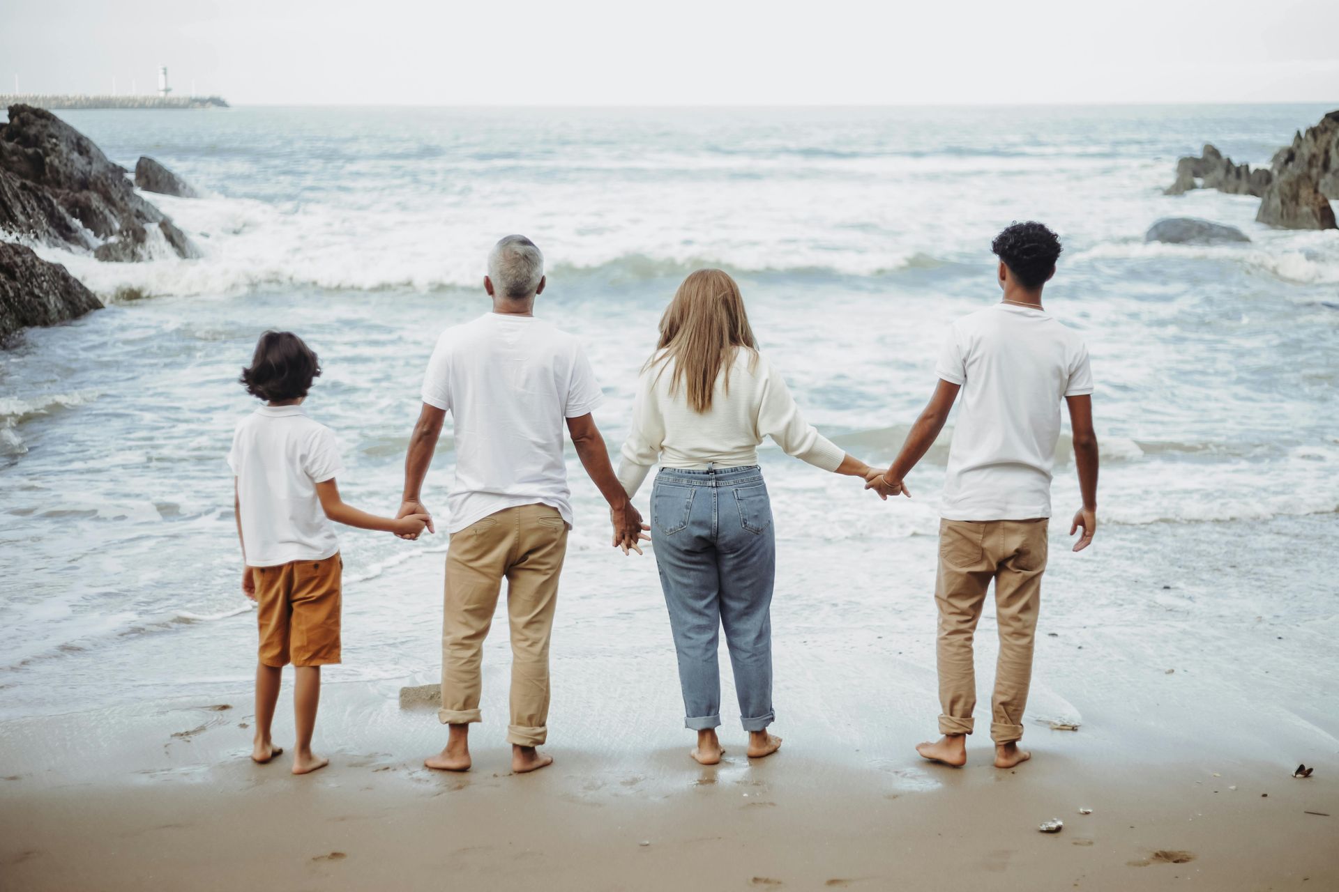 Family holding hands, standing on a beach, facing the ocean. Waves gently lap the shore.